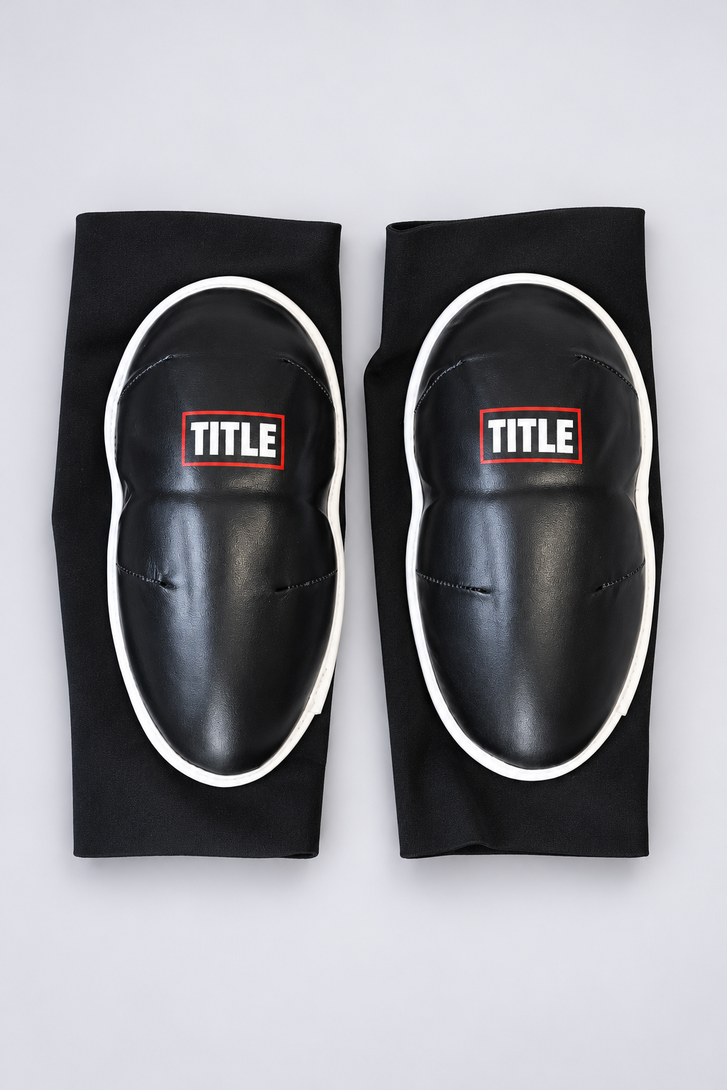 Pair of black Muay Thai elbow pads by TITLE side by side, showing the hinged leather pad and stitched darts