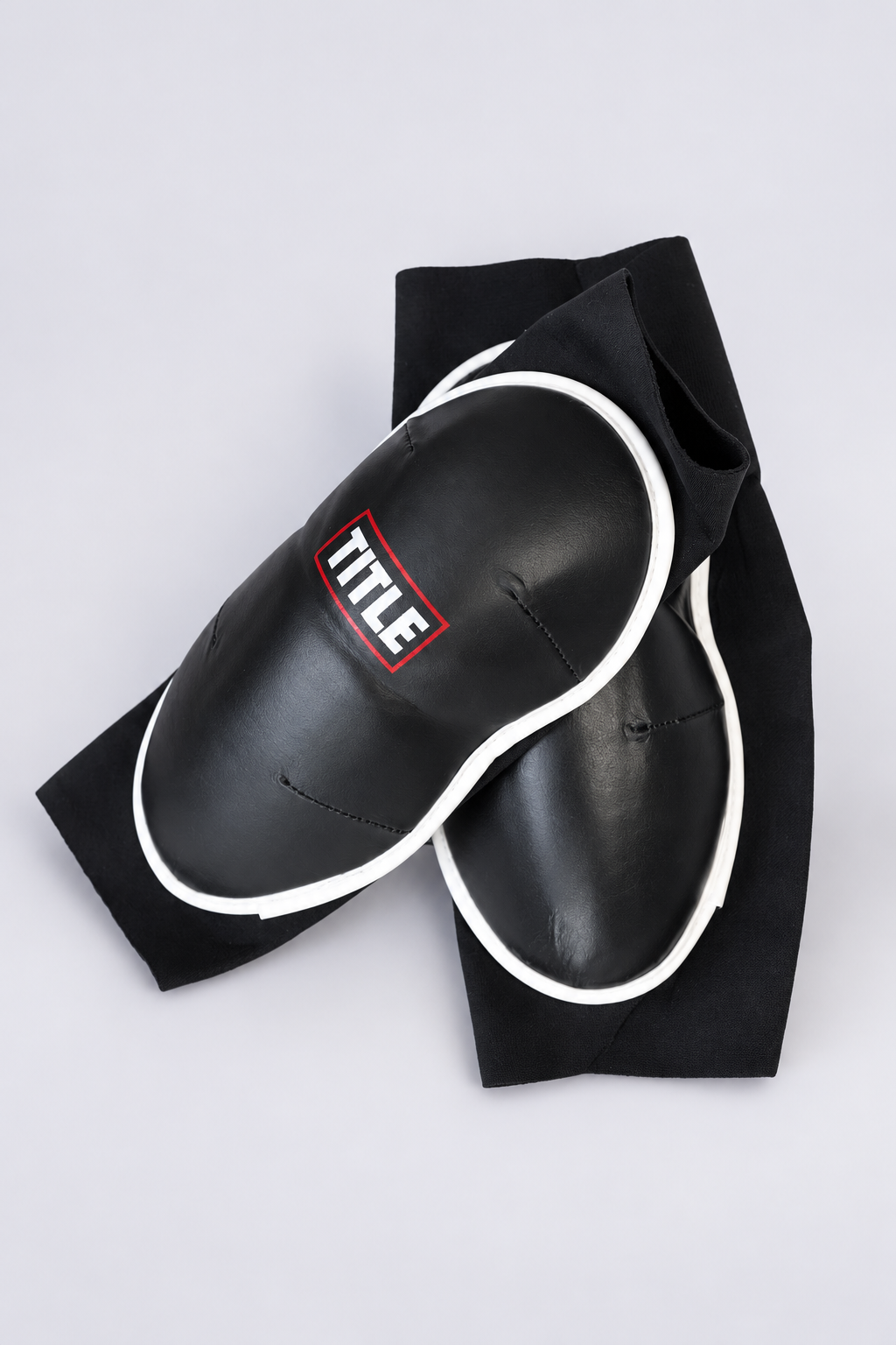 Black slip-on Muay Thai knee pads by TITLE overlapping, top-down view of the leather pad and TITLE logo