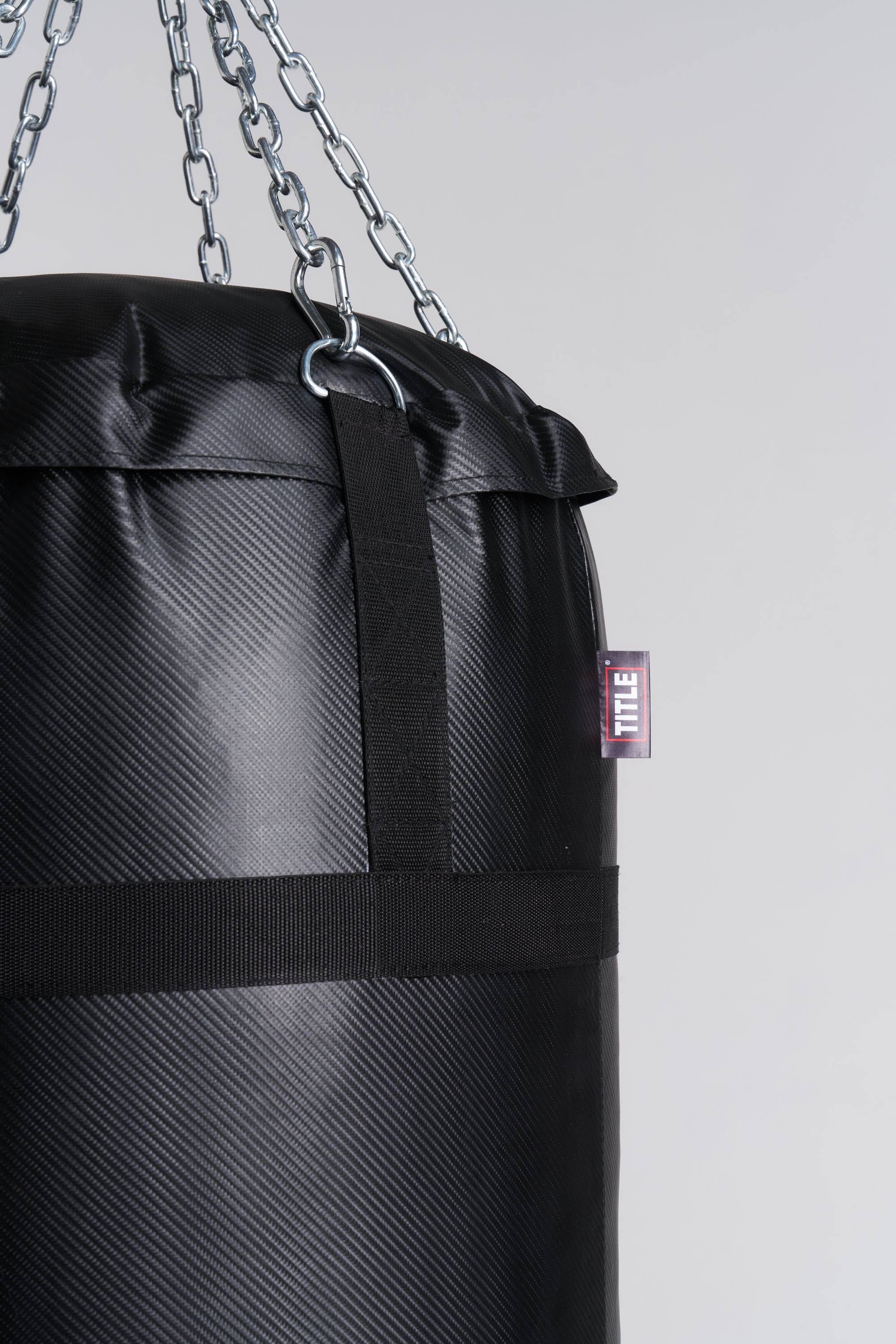 A close-up of a black heavy punching bag with a textured surface, hanging from silver metal chains. A small red and white TITLE brand tag is visible on the side.
