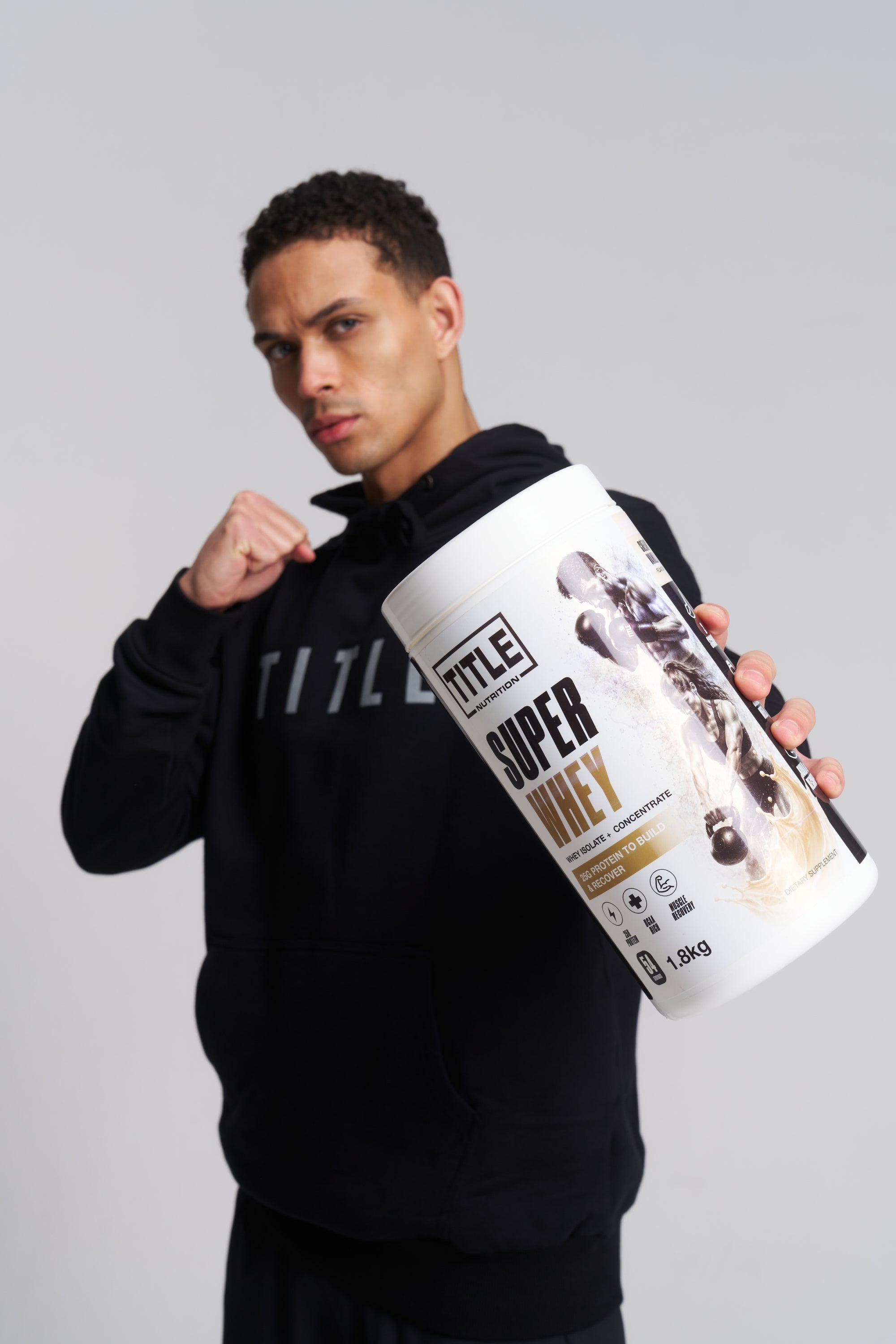 A man in a black hoodie holds a large white tub of TITLE1 Super Whey protein powder against a light gray studio background.