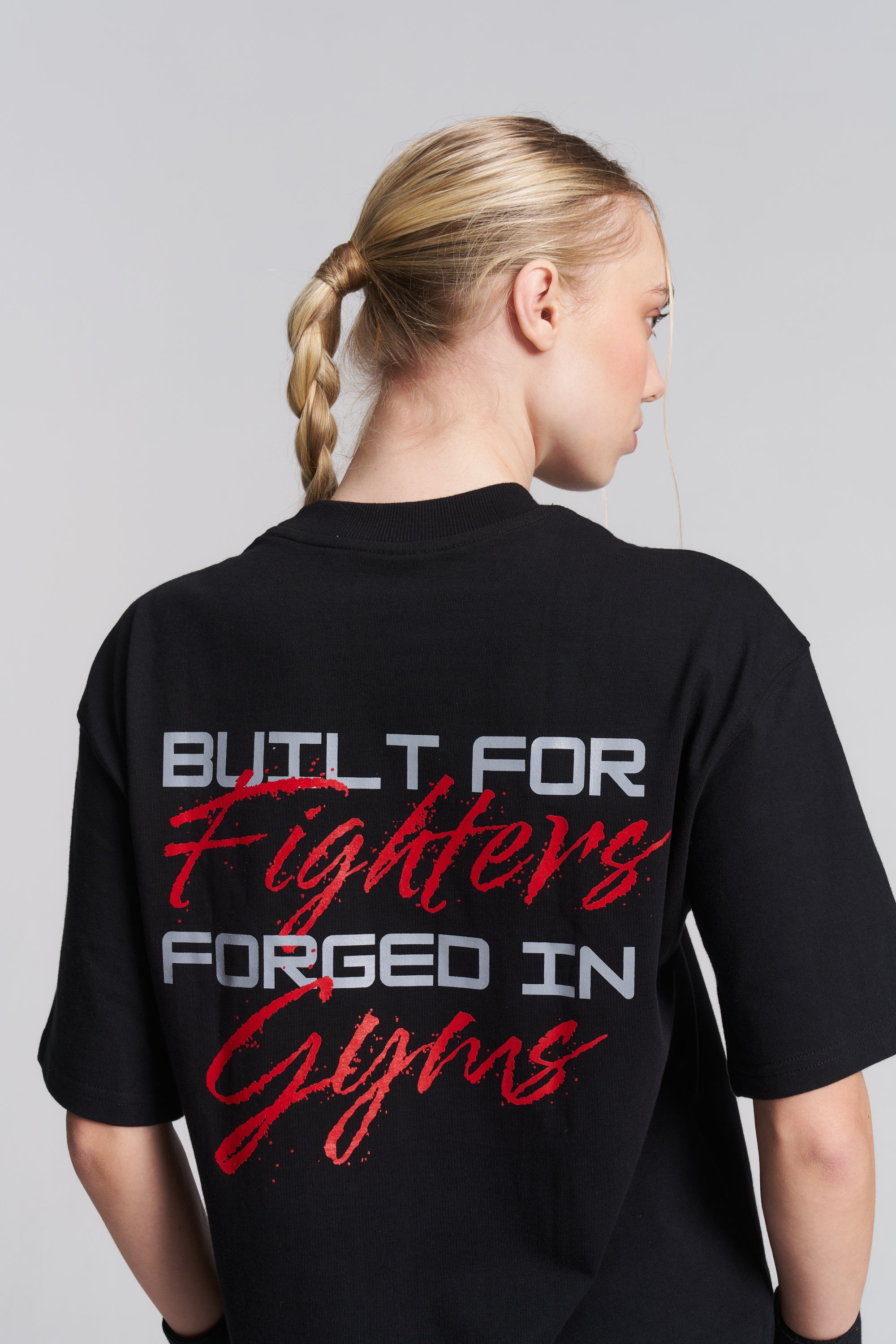 A woman shown from the back wearing a black graphic t-shirt with the text 'BUILT FOR Fighters FORGED IN Gyms' printed on the back in white and red.