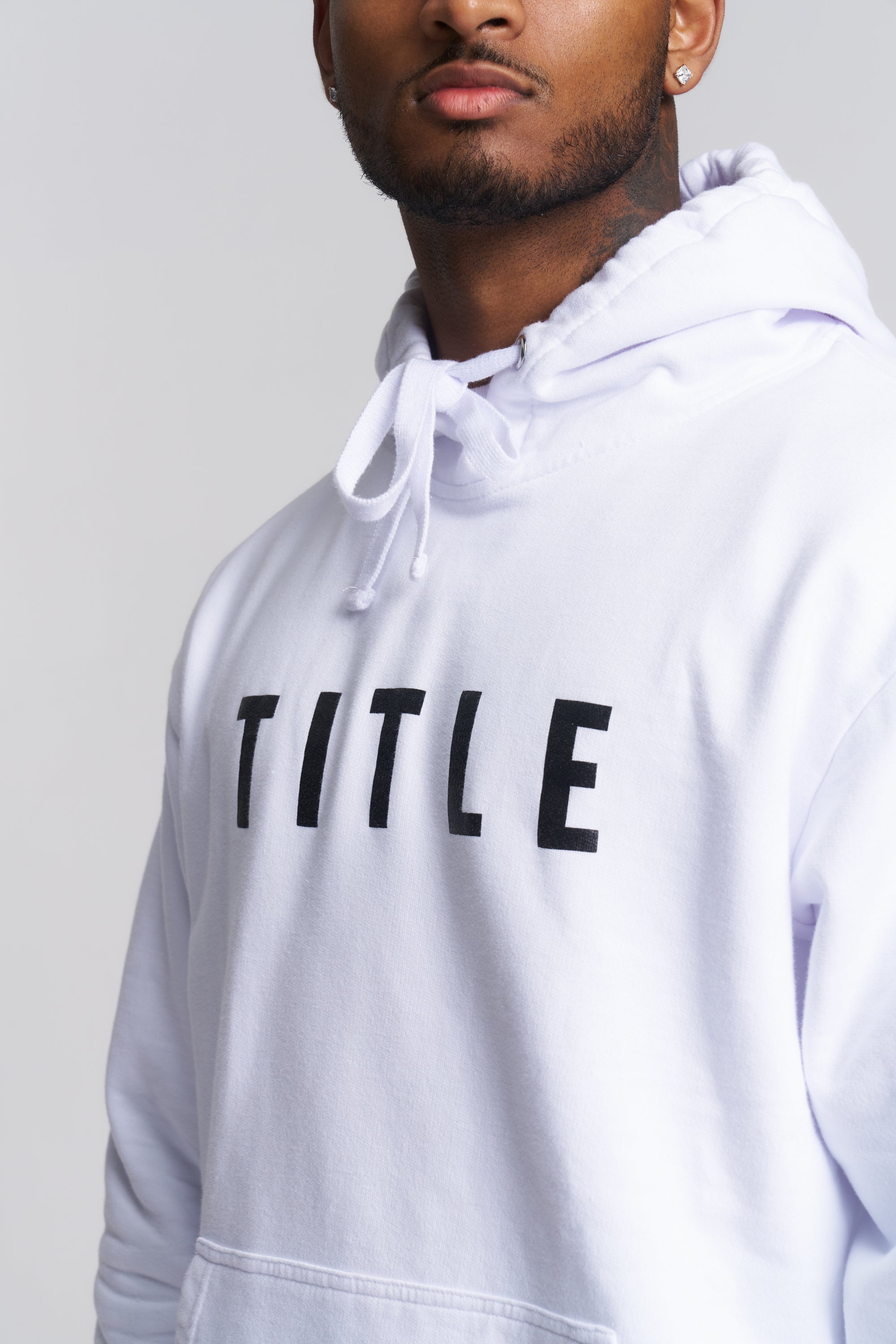 A close-up of a man wearing a white pullover hoodie with the word 'TITLE' printed in bold black letters across the chest, against a light gray background.