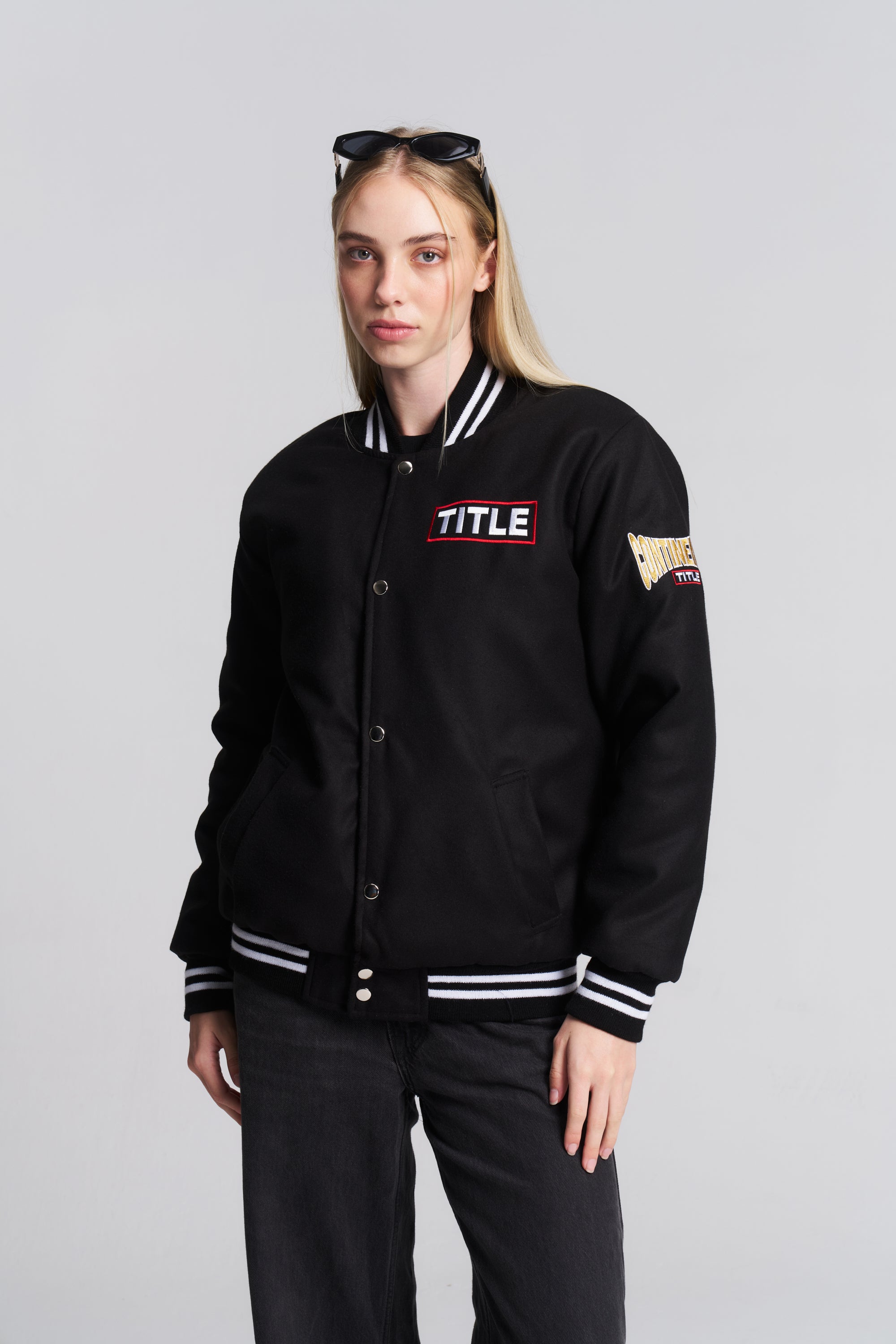 A model wearing a black varsity-style jacket with white and red accents, featuring 'TITLE' embroidered on the chest and a patch on the sleeve.