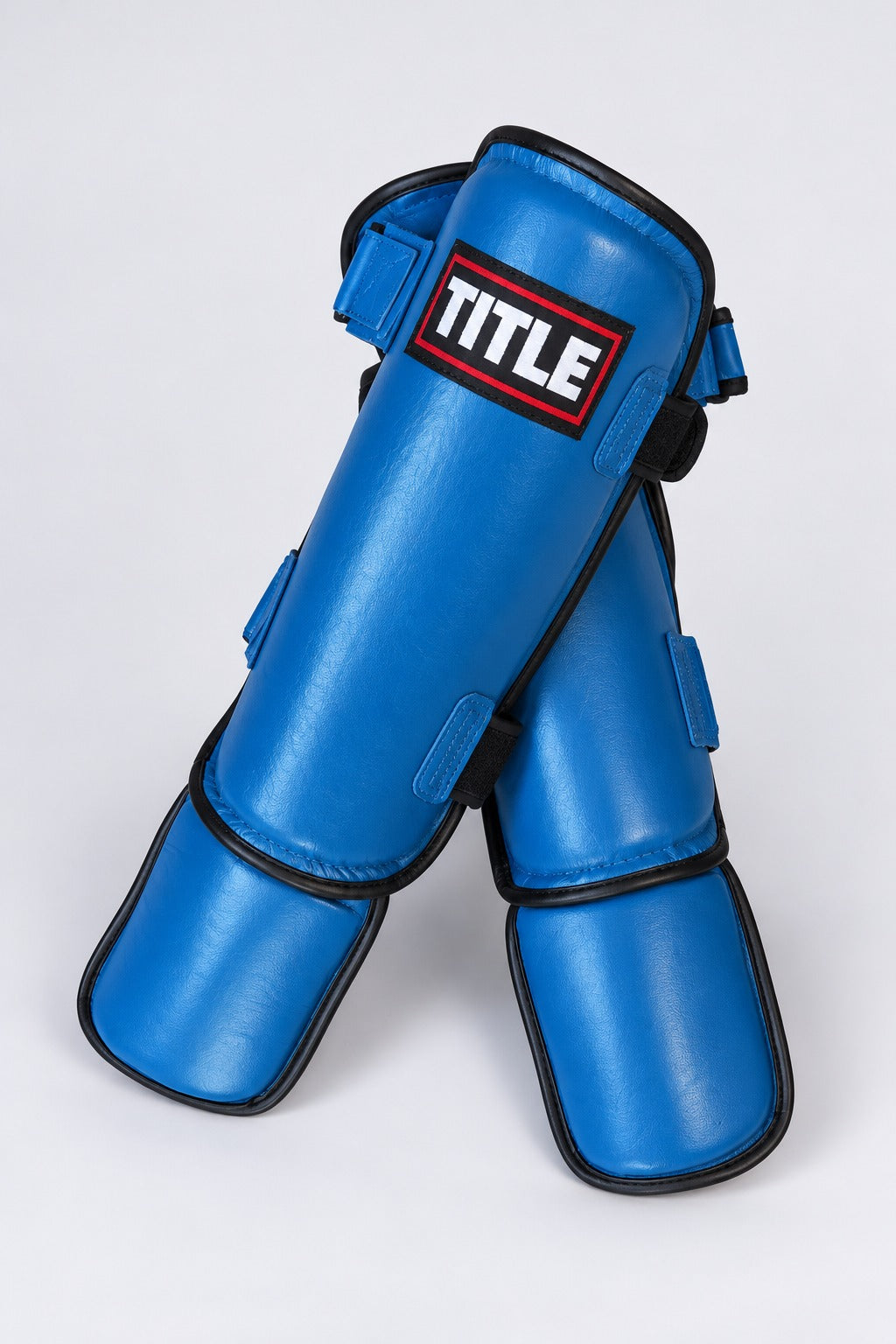 Pair of blue leather kickboxing shin guards by TITLE crossed at an angle, showing strap keepers and instep