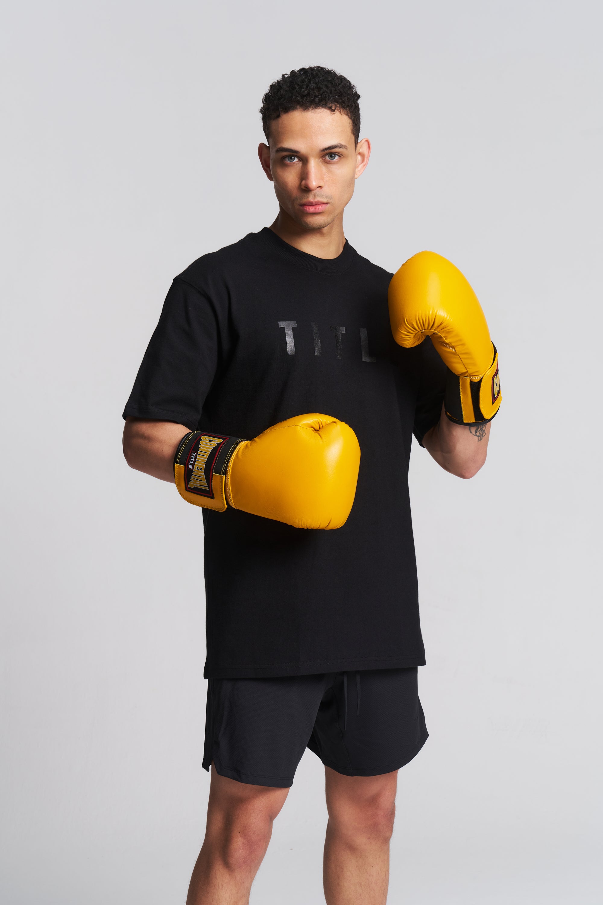 A man wearing a black graphic t-shirt, black athletic shorts, and bright yellow boxing gloves, posing in a fighting stance against a light gray studio background.
