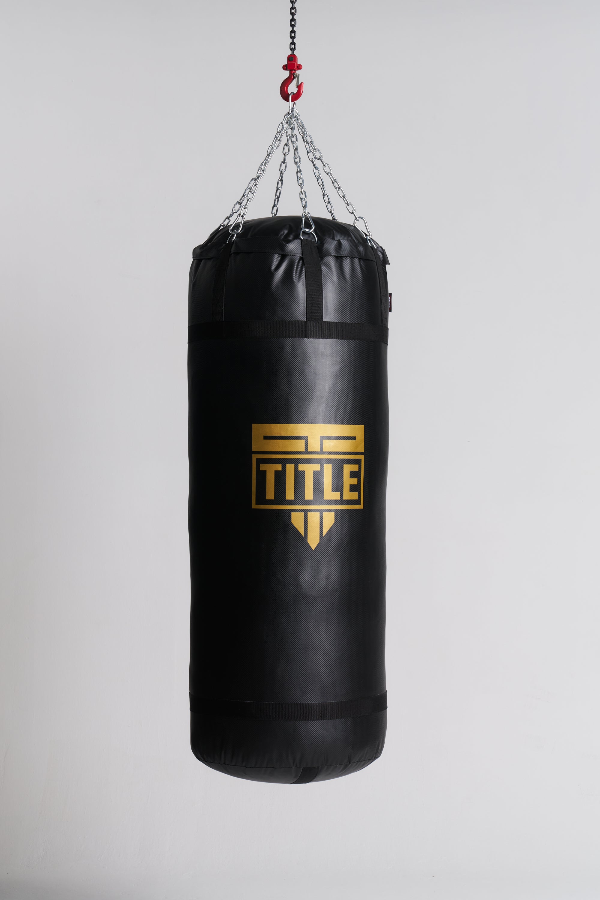A black heavy punching bag hanging from a chain with a red hook, featuring a gold TITLE logo on the front, set against a plain light gray background.