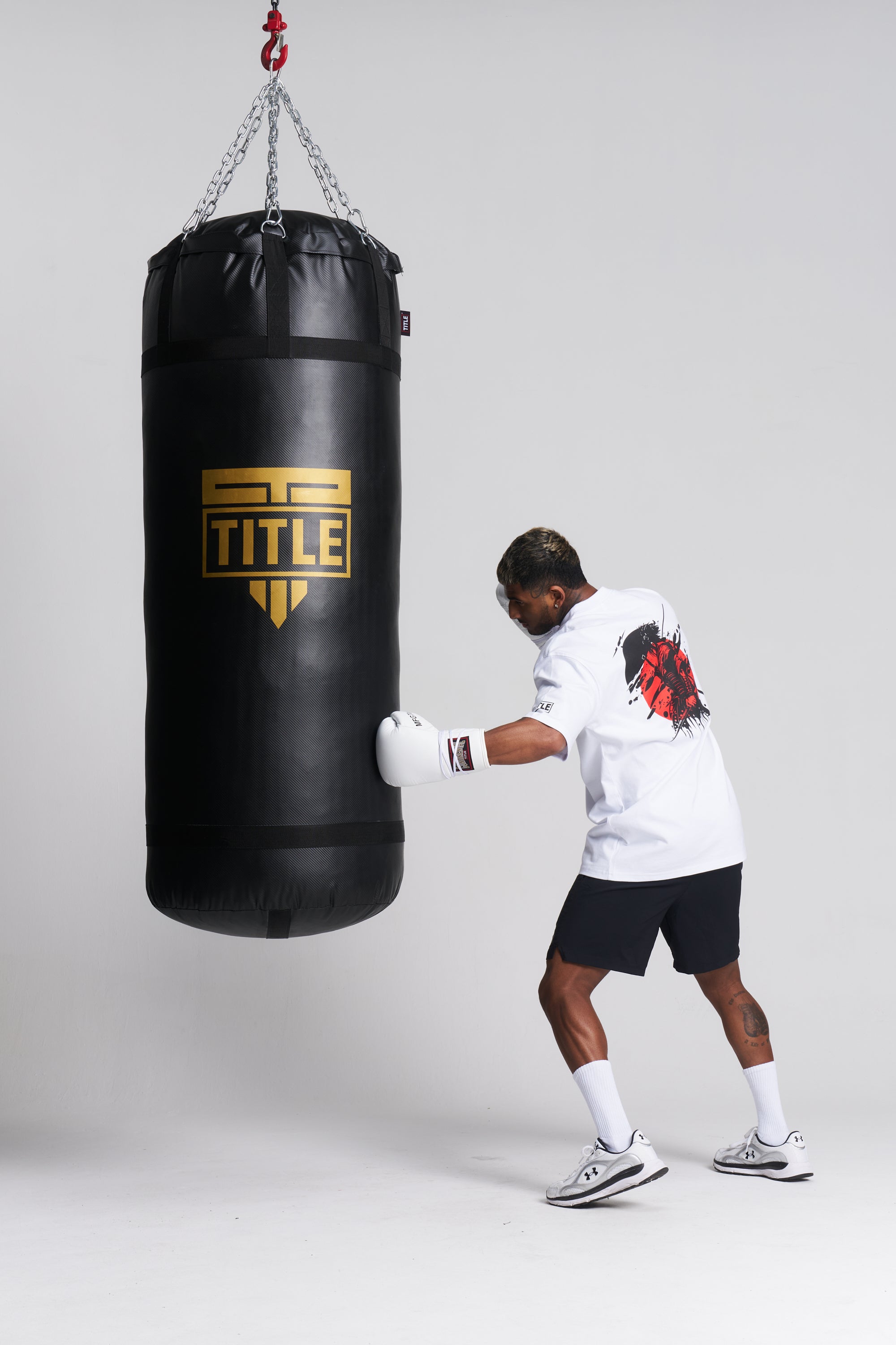 A man in a white t-shirt, black shorts, and boxing gloves punches a black heavy bag hanging from chains. The bag features a gold and black TITLE logo.