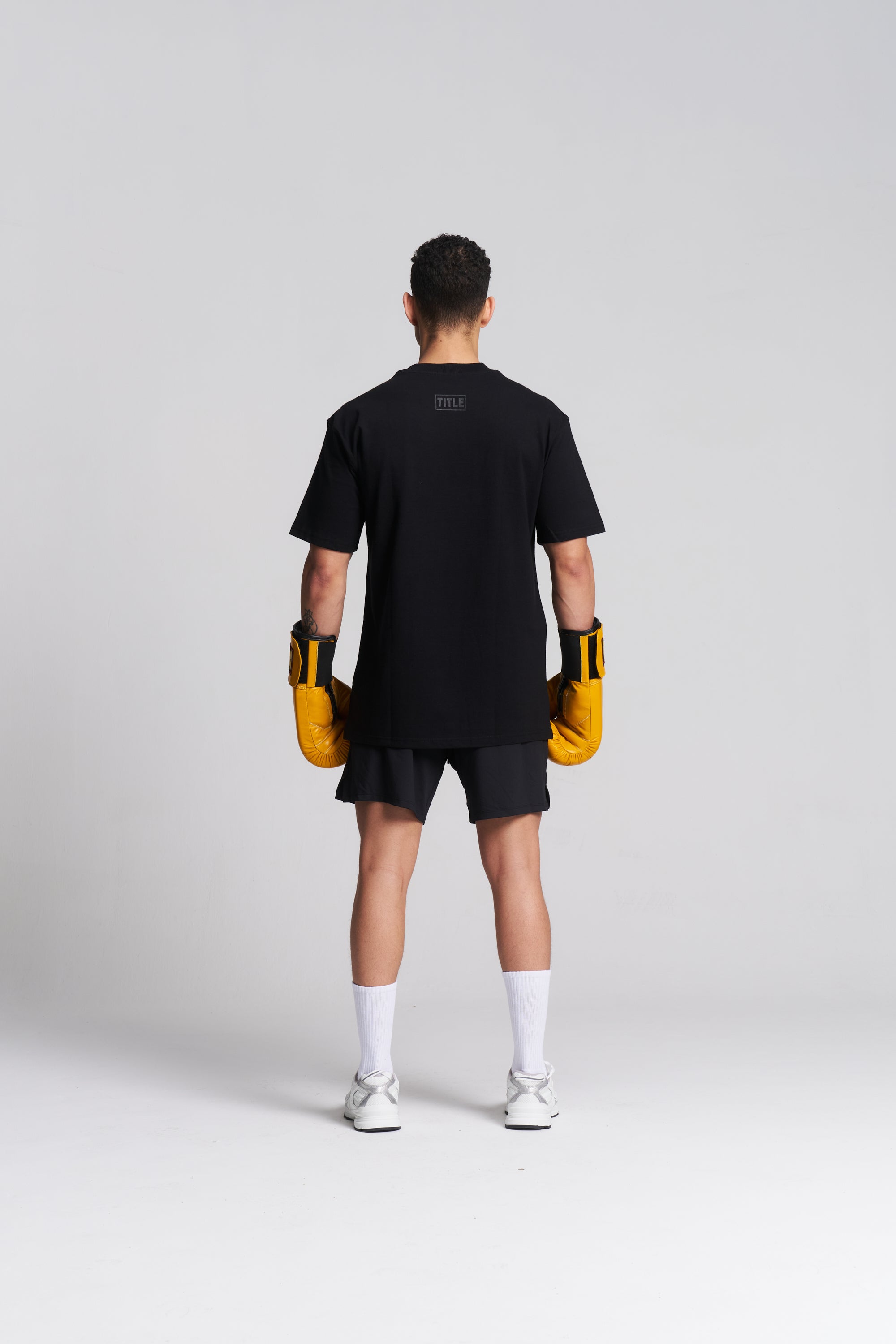 A man is shown from the back wearing a black t-shirt, black shorts, white socks, and white sneakers, while holding yellow boxing gloves against a light gray studio background.