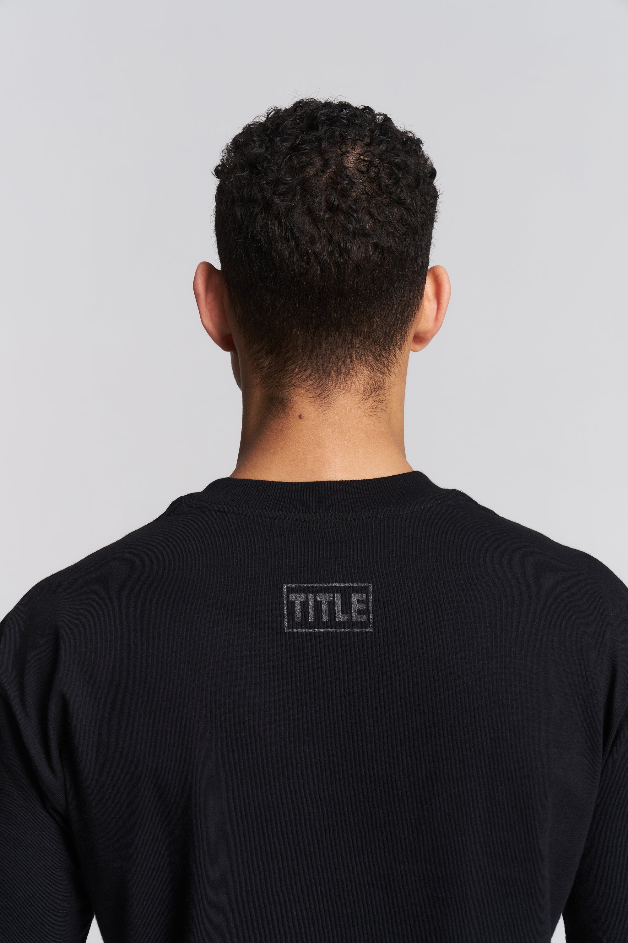 Back view of a man wearing a black long-sleeve crewneck sweatshirt with a tonal black 'TITLE' logo box on the upper back, set against a light gray studio background.