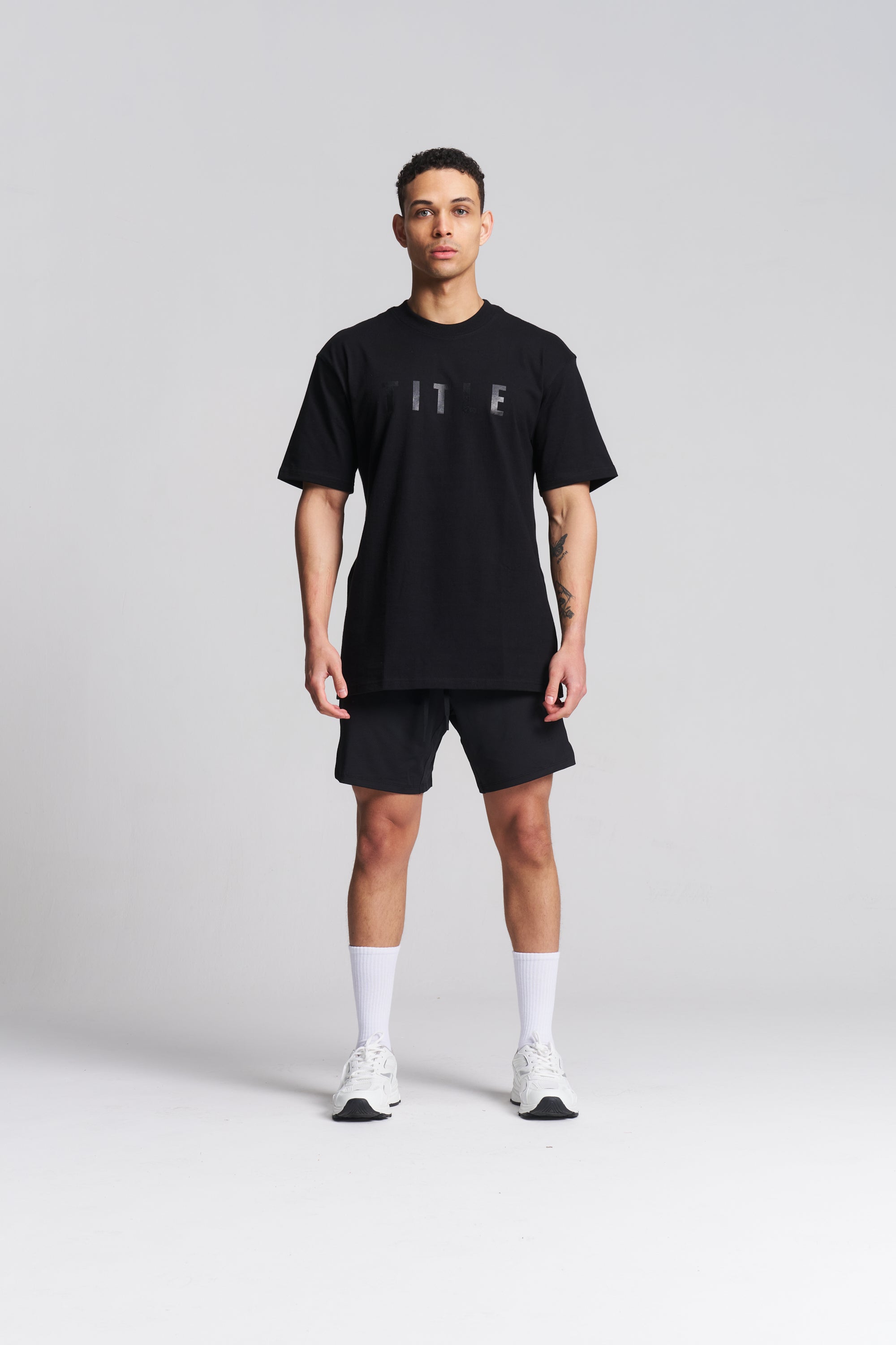 A man modeling a black short-sleeve t-shirt with a subtle gray 'ITF' logo on the chest, paired with black athletic shorts, white socks, and white sneakers, against a light gray studio background.