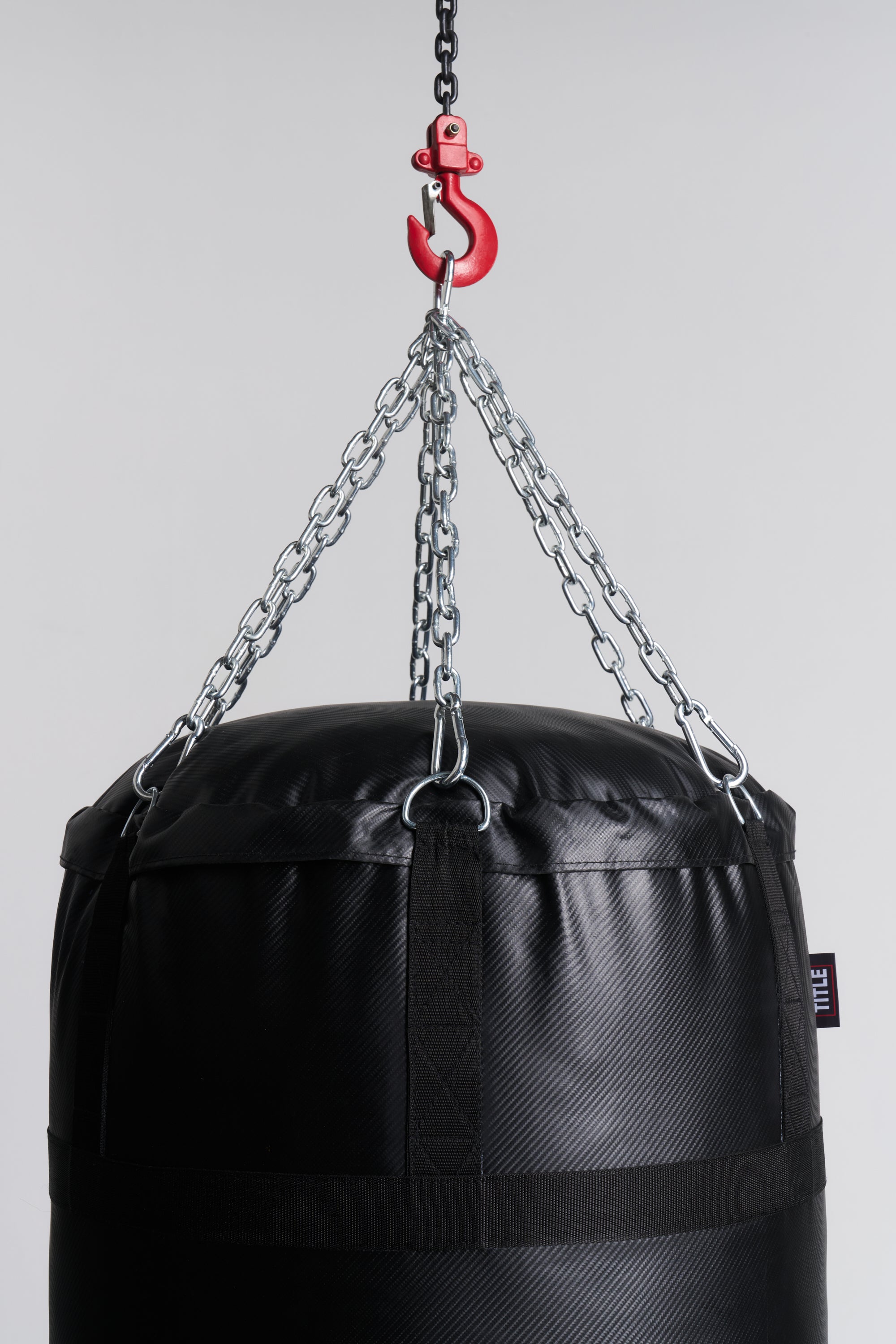 A black heavy punching bag suspended by silver metal chains from a red hook on a light gray background.
