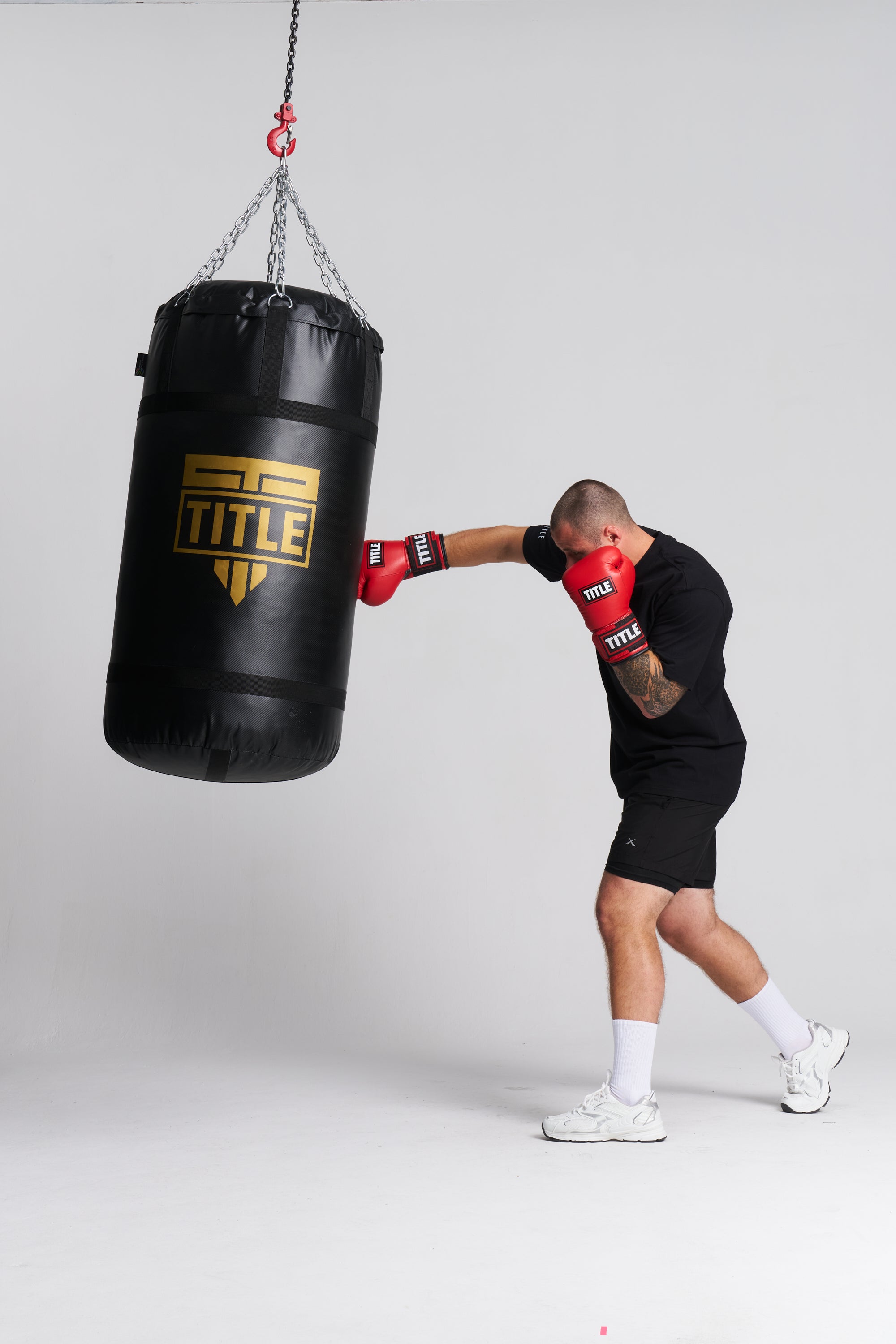 A man in a black athletic outfit and red boxing gloves punches a black Title brand hanging heavy bag against a light gray studio background.