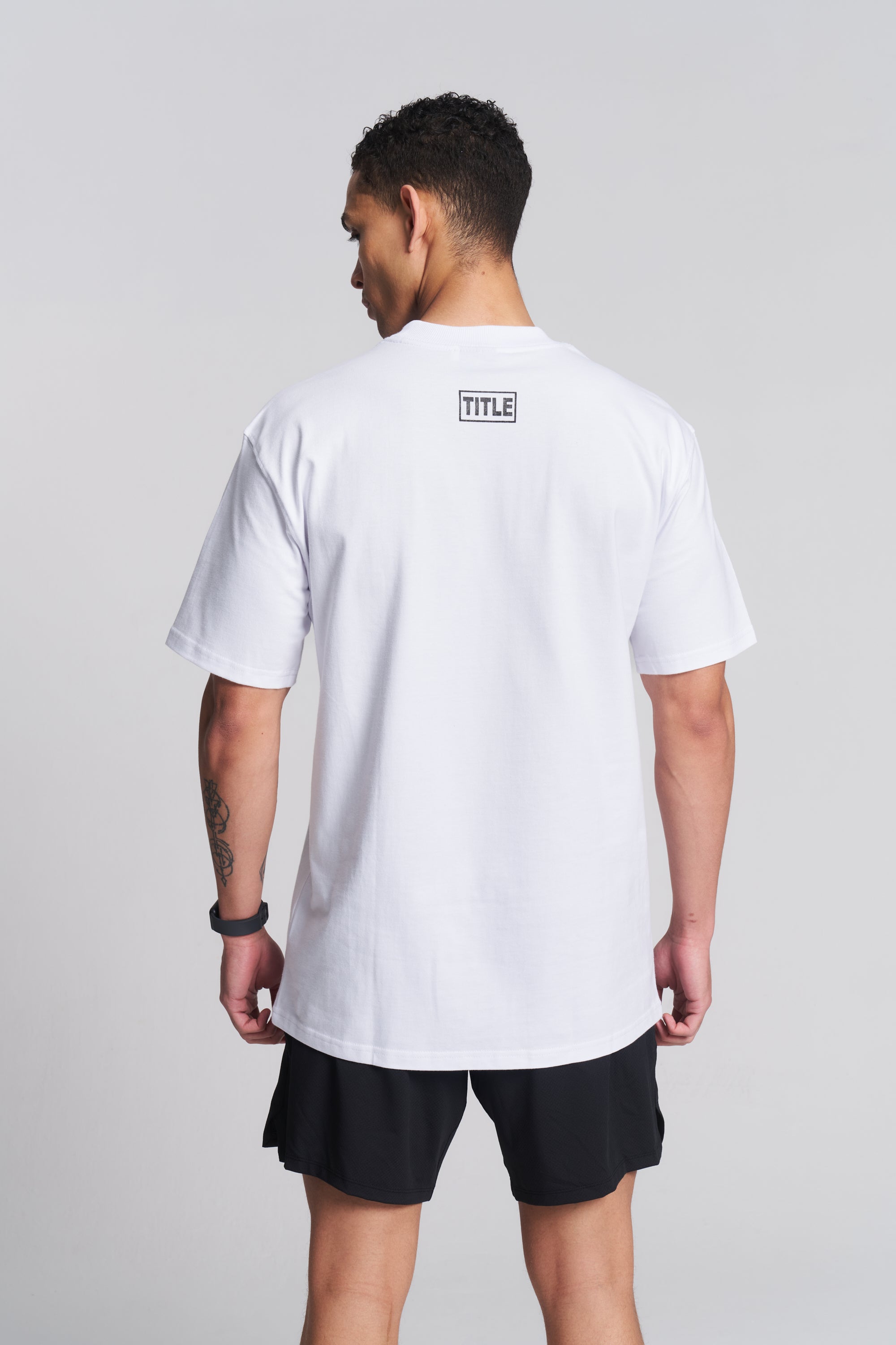 A man viewed from the back wearing a white short-sleeve T-shirt with a small black rectangular logo patch on the upper back that reads TITLE, paired with black shorts, against a light gray studio background.