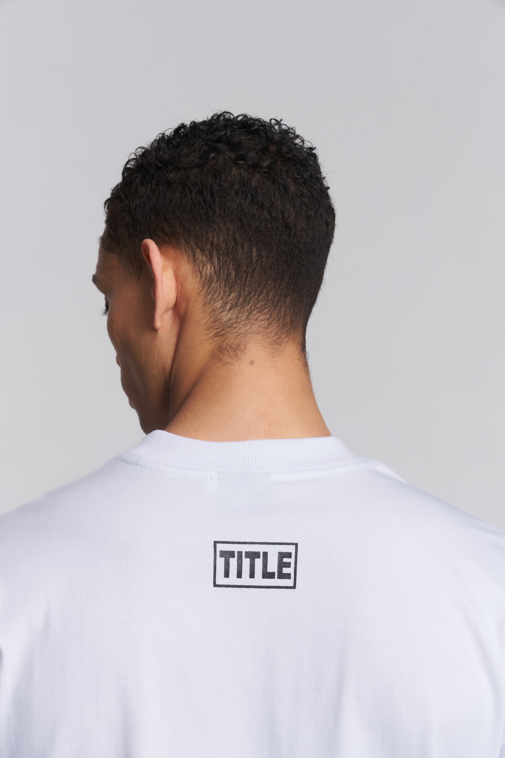 Back view of a man wearing a white crewneck t-shirt with a black rectangular 'TITLE' logo printed on the upper back, set against a plain light gray background.