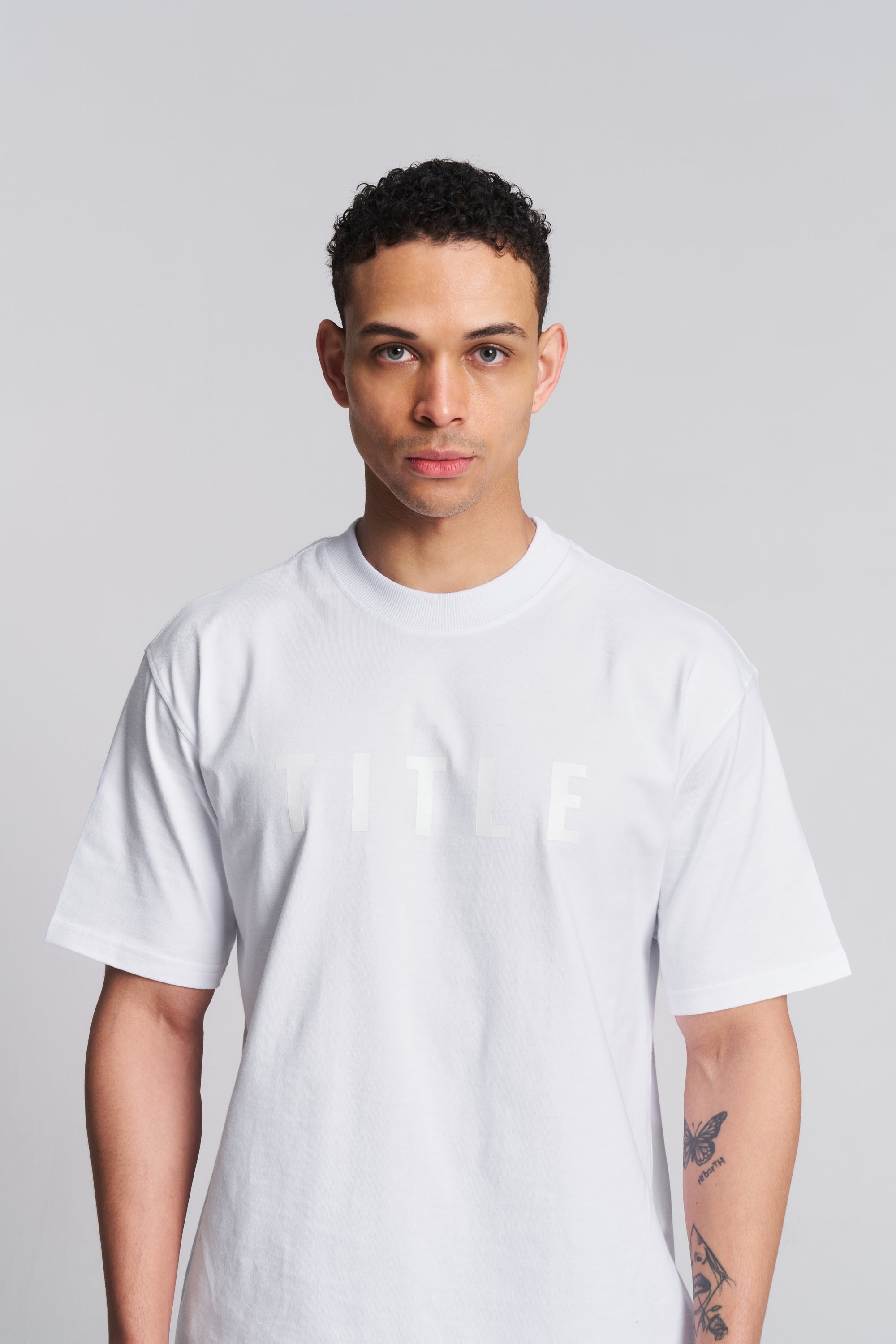 A man wearing a white short-sleeve crewneck t-shirt with a subtle, tonal 'TITLE' logo printed across the chest, photographed against a solid light gray background.