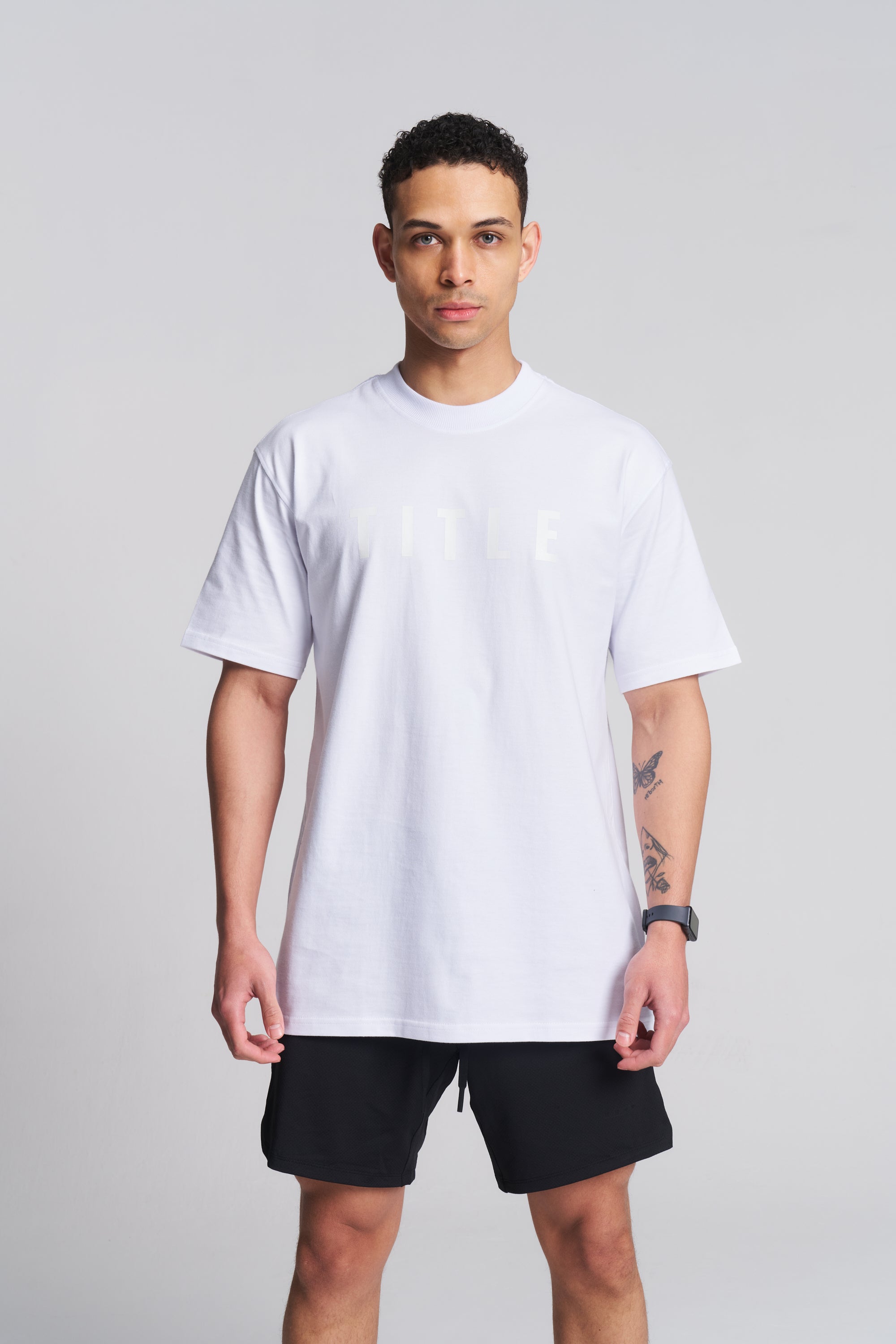 A man modeling a white short-sleeve crewneck t-shirt with a subtle tonal graphic on the chest, paired with black athletic shorts, against a solid light gray background.