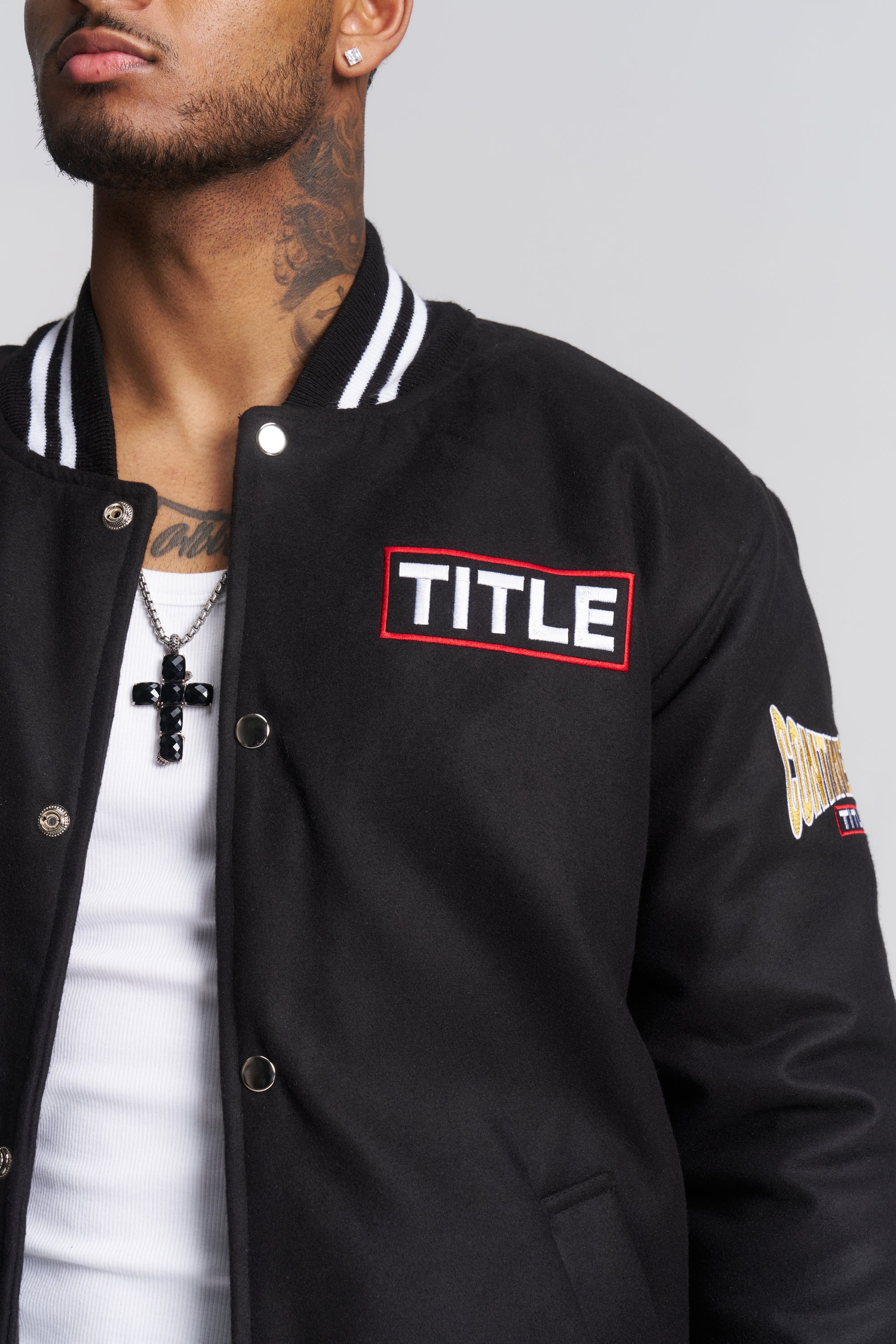 A close-up of a man wearing a black varsity-style jacket with a prominent red and white 'TITLE' patch on the chest.