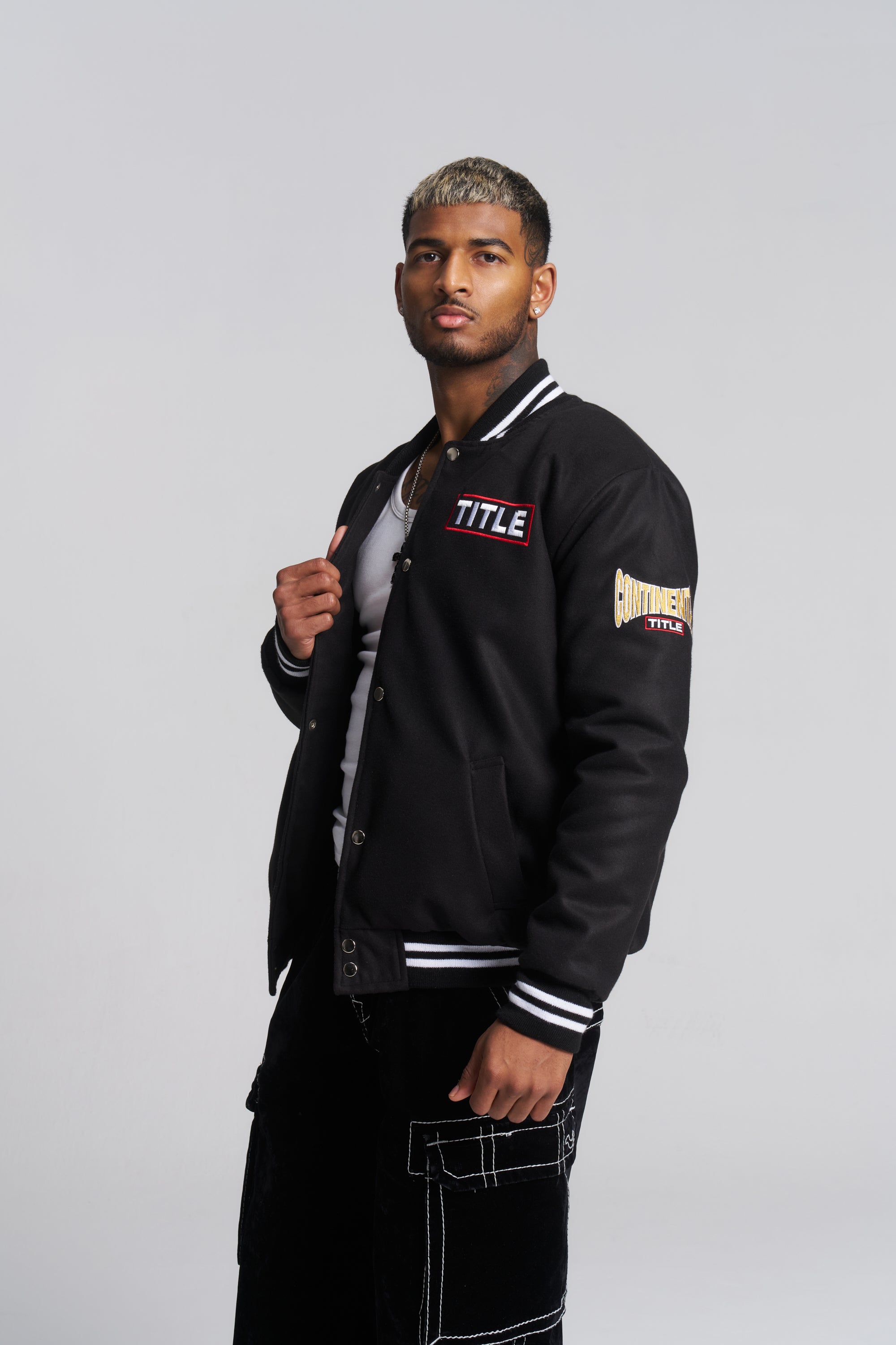 A man wearing a black varsity-style jacket with white striped ribbed trim, featuring a 'TITLE' logo on the chest and a sleeve patch, paired with black pants.