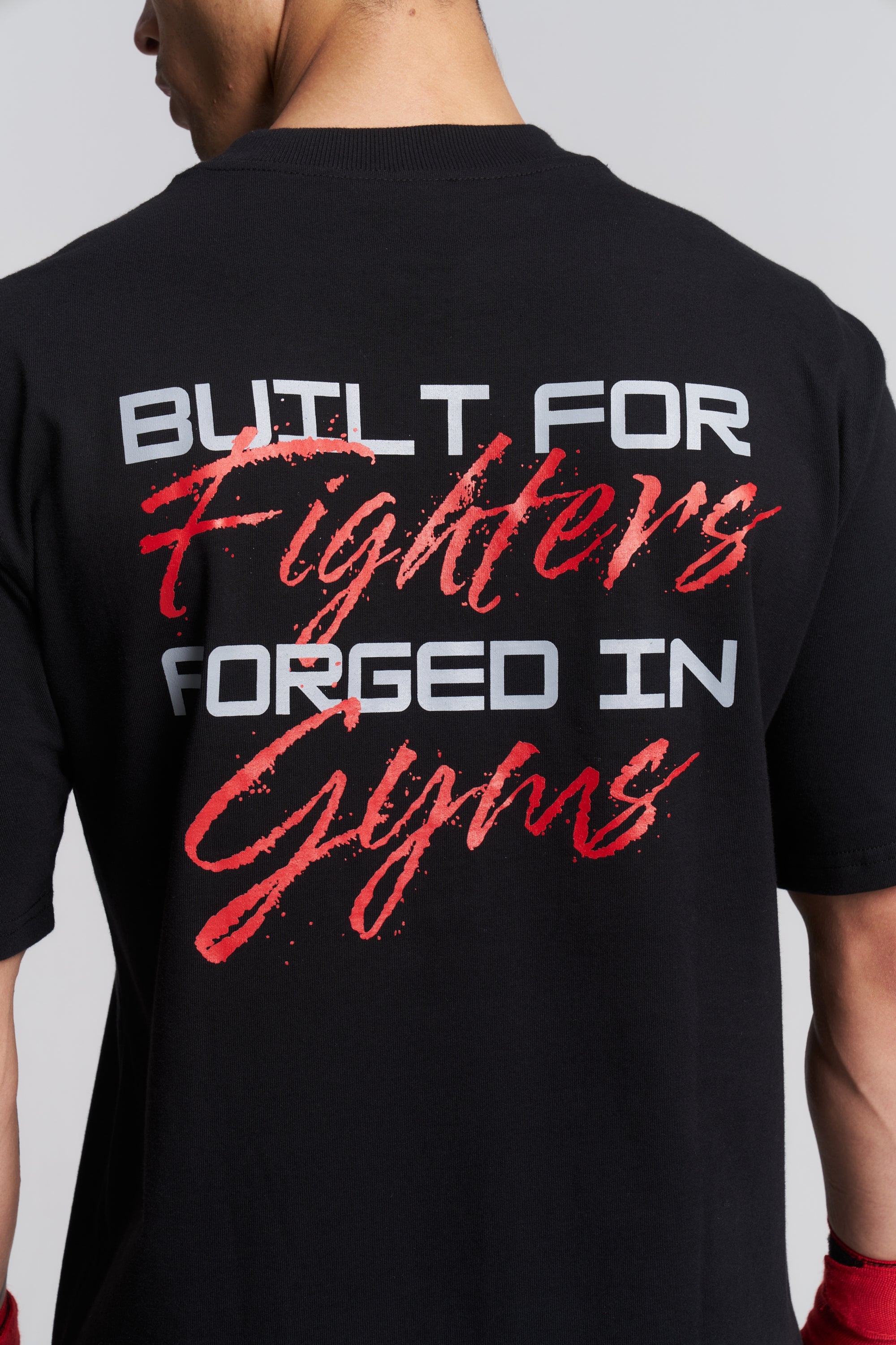 A close-up back view of a person wearing a black t-shirt with a large graphic print on the back. The text reads 'BUTLT FOR Fighters FORGED IN Gyms' in white and red lettering, set against a plain light gray background.