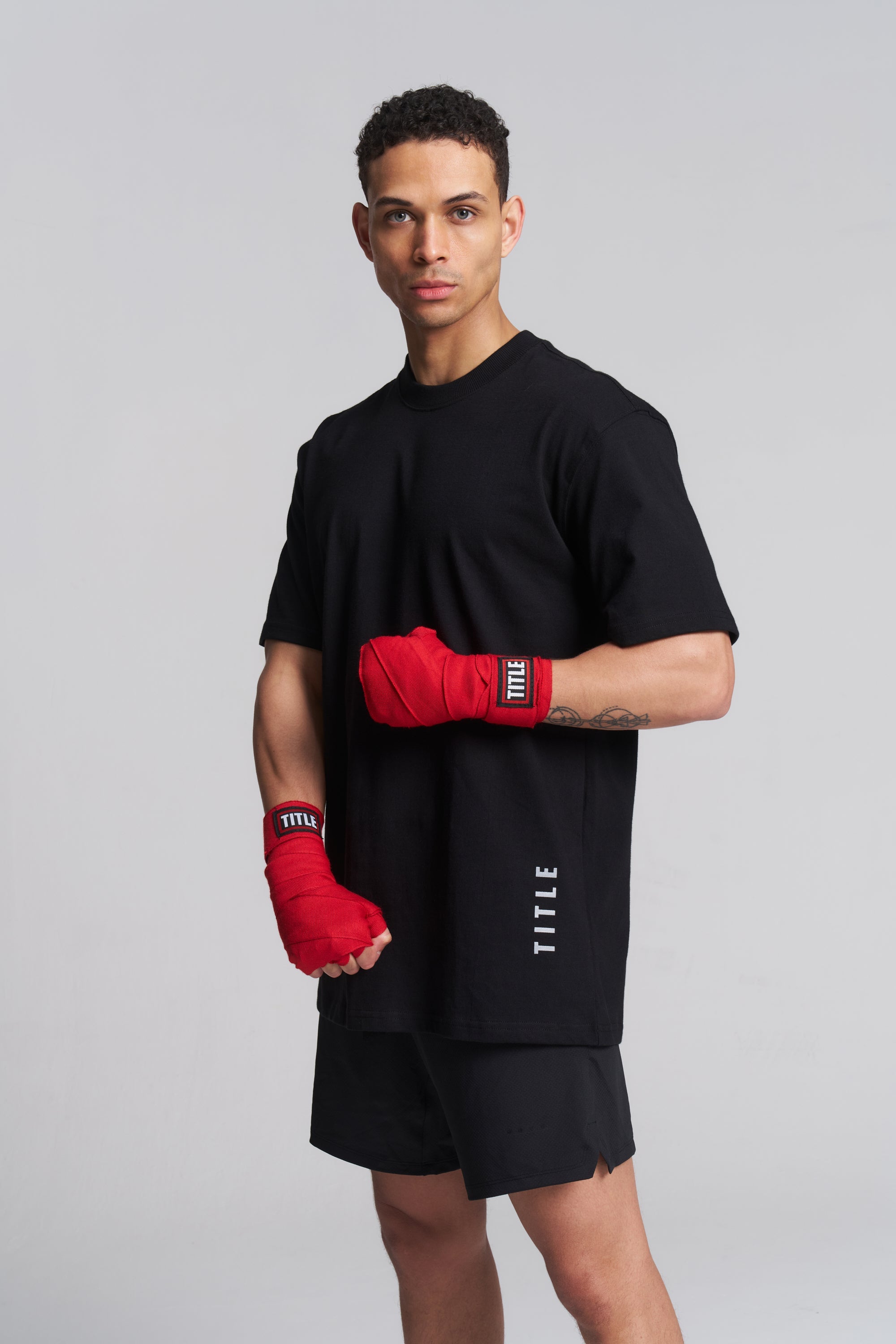 A man modeling a black short-sleeve athletic t-shirt and matching black shorts, with red boxing wraps on his hands and forearms. The t-shirt features a vertical white 'TITLE' logo on the lower right side.