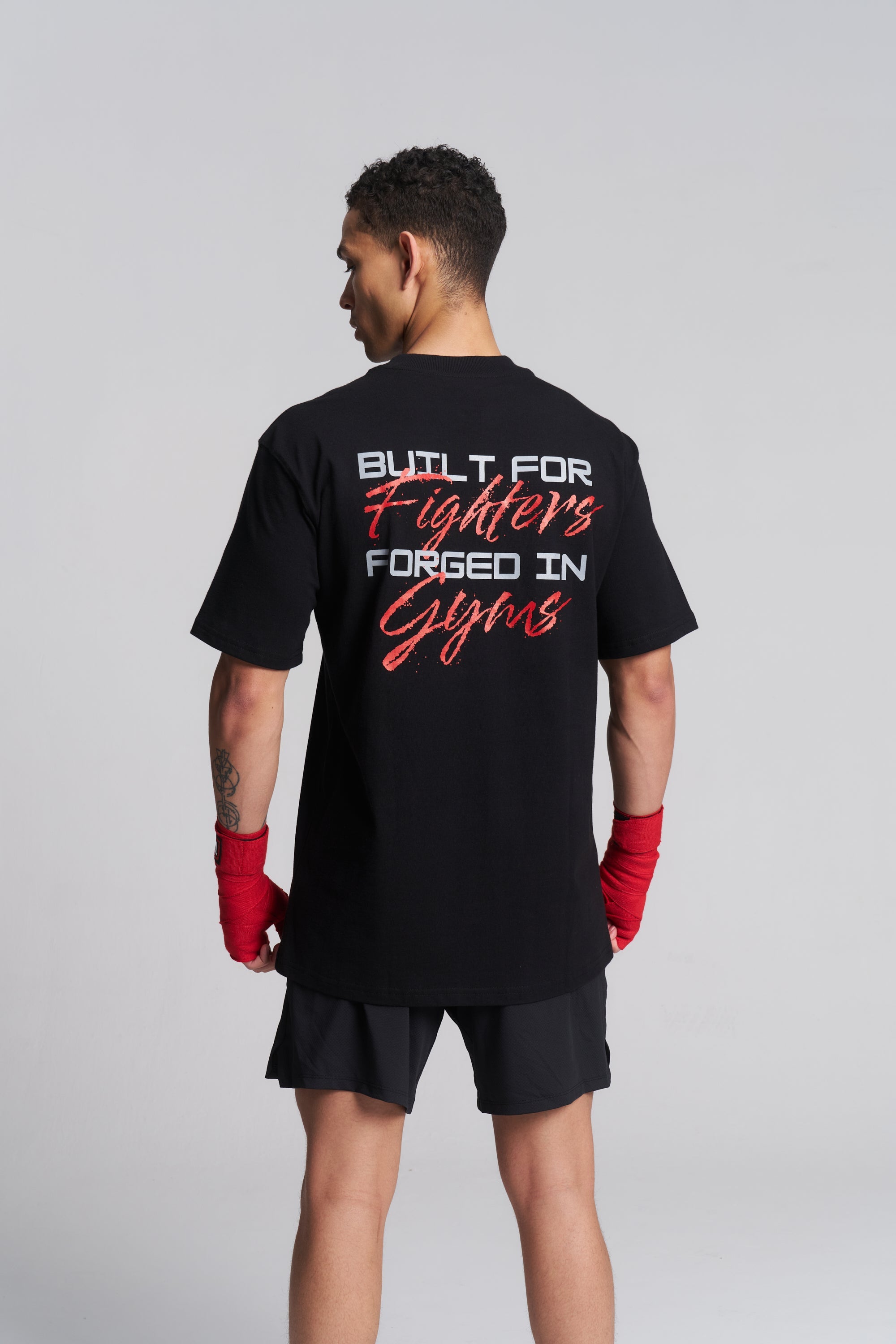 A man viewed from the back wearing a black graphic t-shirt with 'BUILT FOR Fighters FORGED IN Gyms' printed in white and red on the back, paired with black shorts and red boxing gloves, against a light gray studio background.