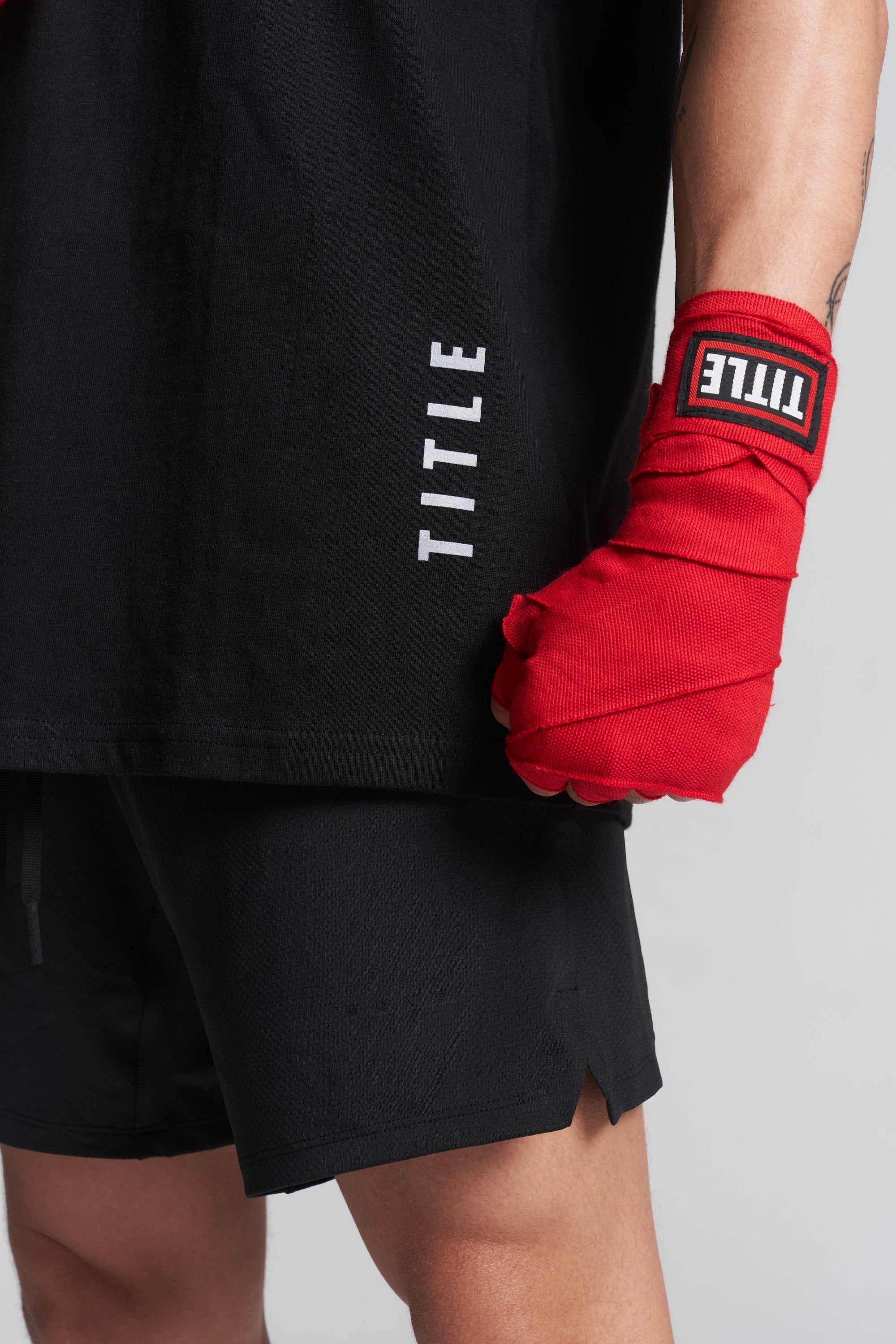 A close-up of a person wearing a black TITLE tank top and black athletic shorts, with a bright red hand wrap on one hand. The TITLE logo is visible on the sleeve of the shirt and on the hand wrap.