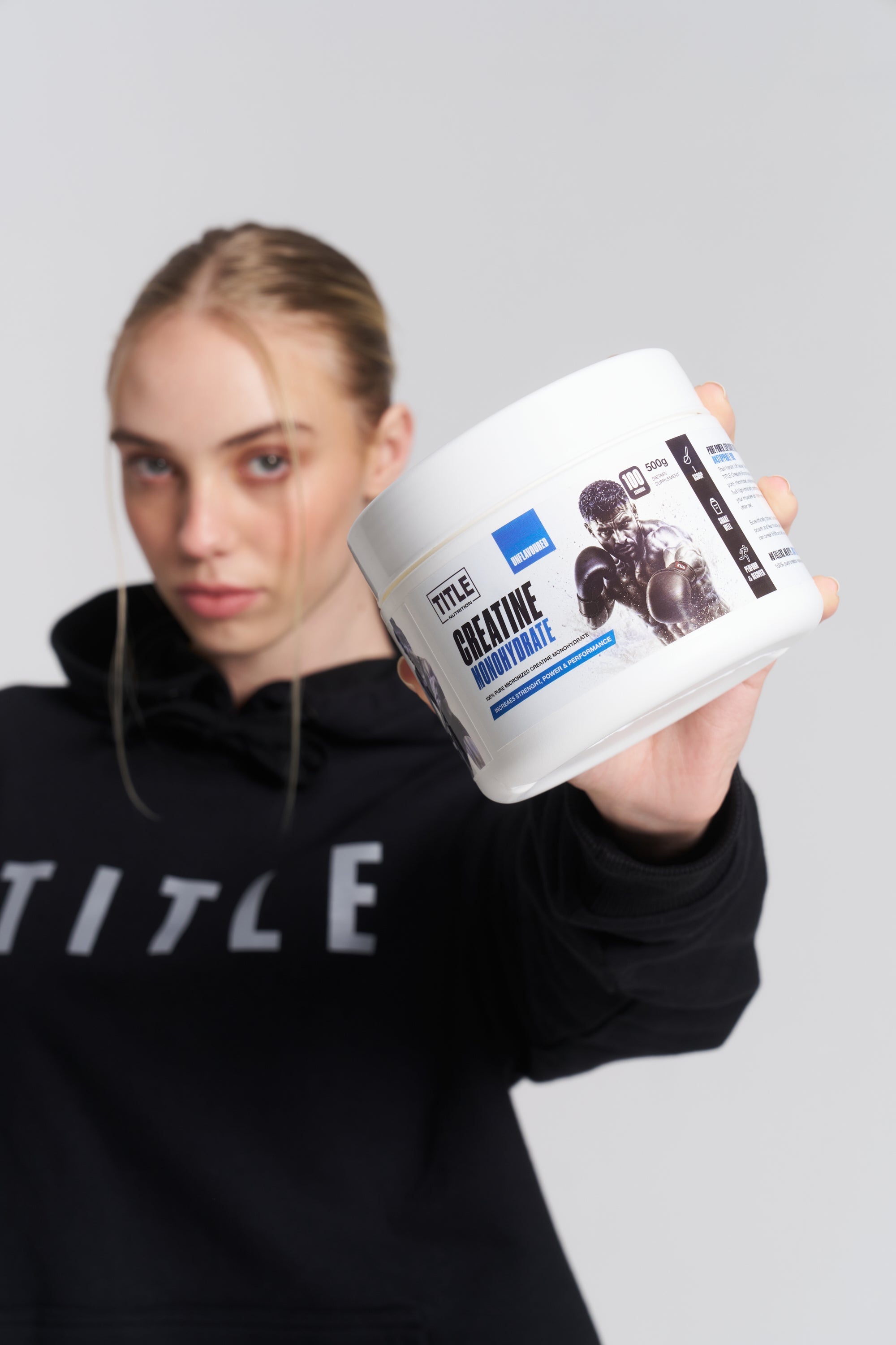 A woman in a black hoodie holding a white tub of TITLE Boxing South Africa 100% micronised creatine monohydrate.
