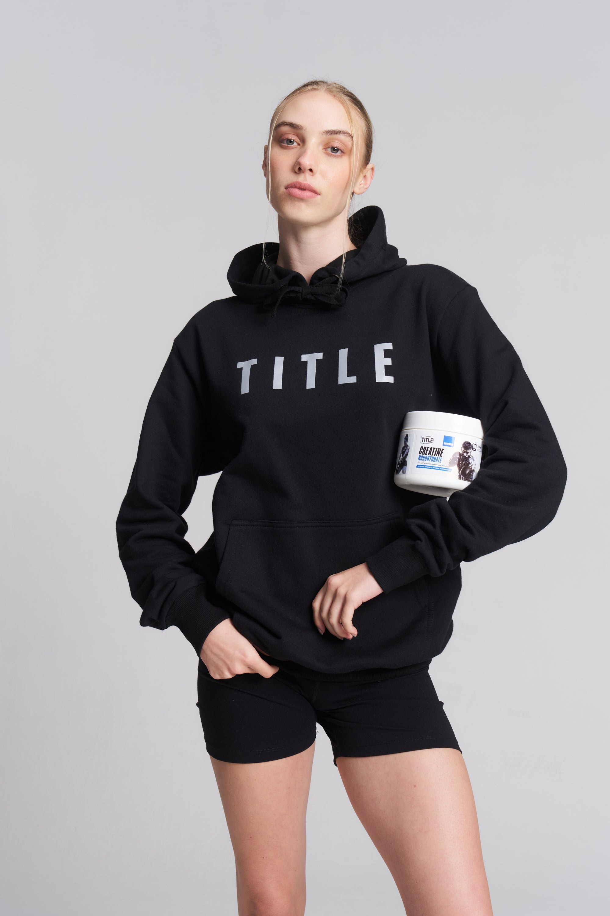A woman wearing a black TITLE Boxing South Africa hoodie holding a tub of TITLE Creatine Monohydrate supplement.