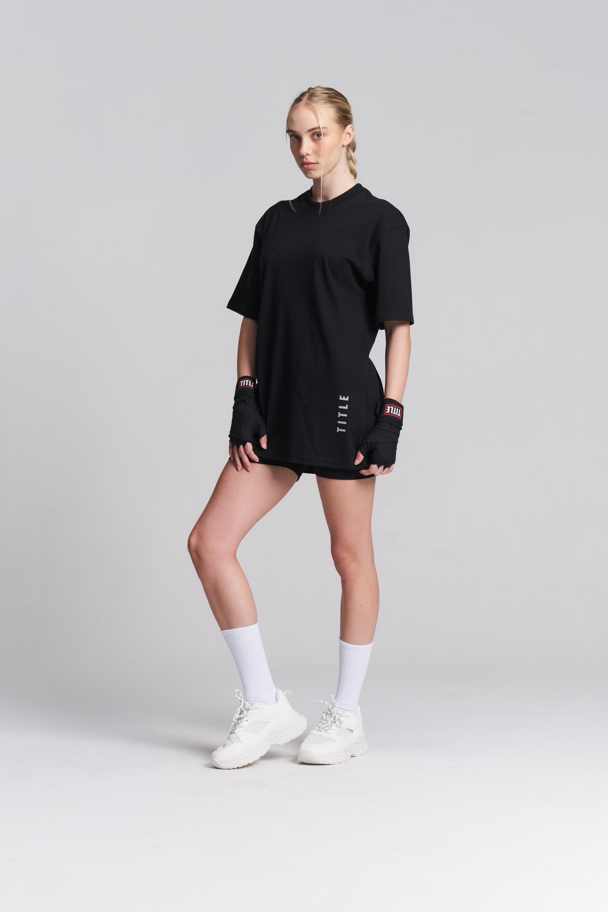 A woman modeling a black oversized short-sleeve athletic top with a high mock neck, paired with black fingerless gloves, white socks, and white sneakers, against a light gray studio background.