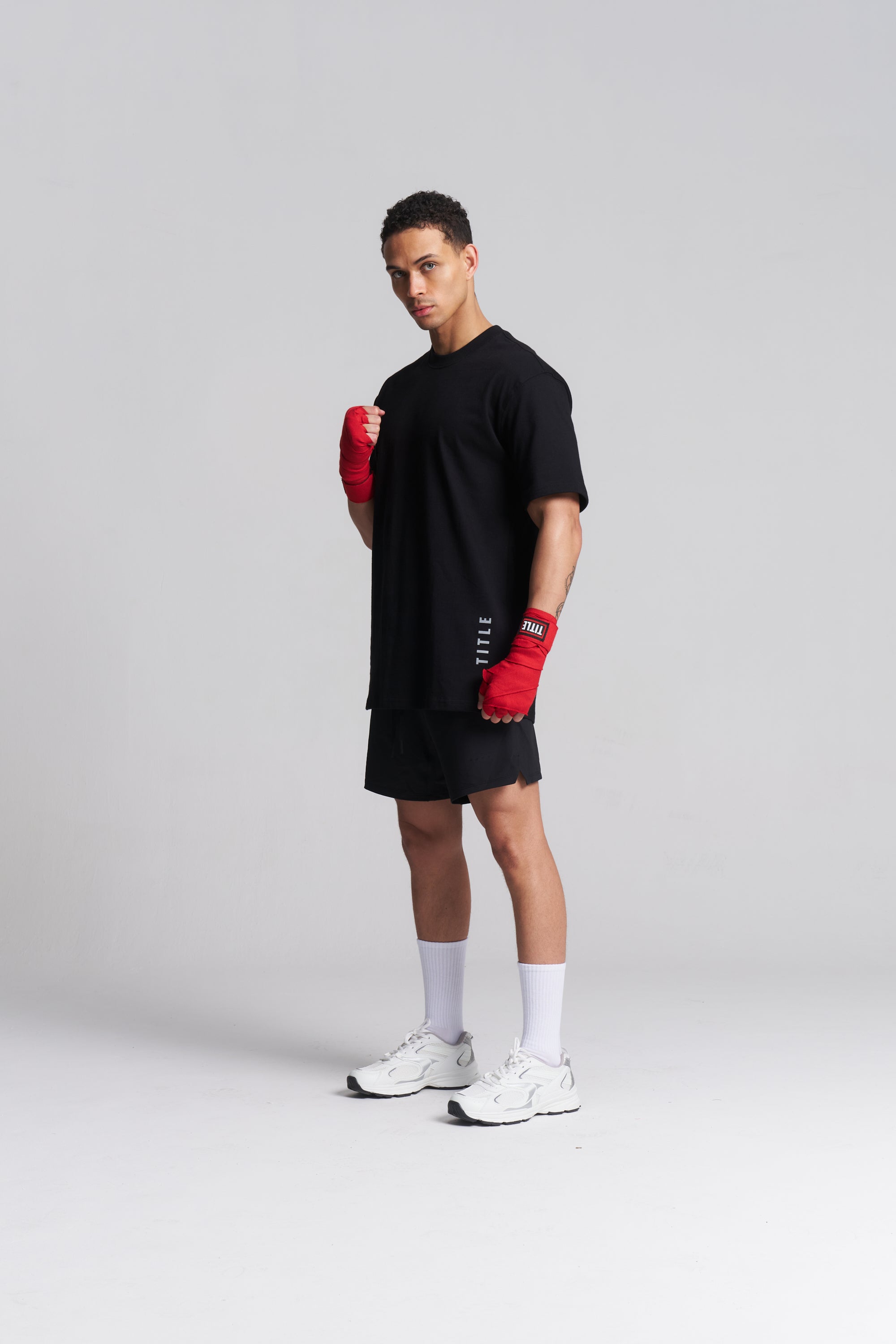 A man modeling a black oversized t-shirt, black athletic shorts, white socks, and white sneakers, while wearing red boxing gloves against a light gray studio background.