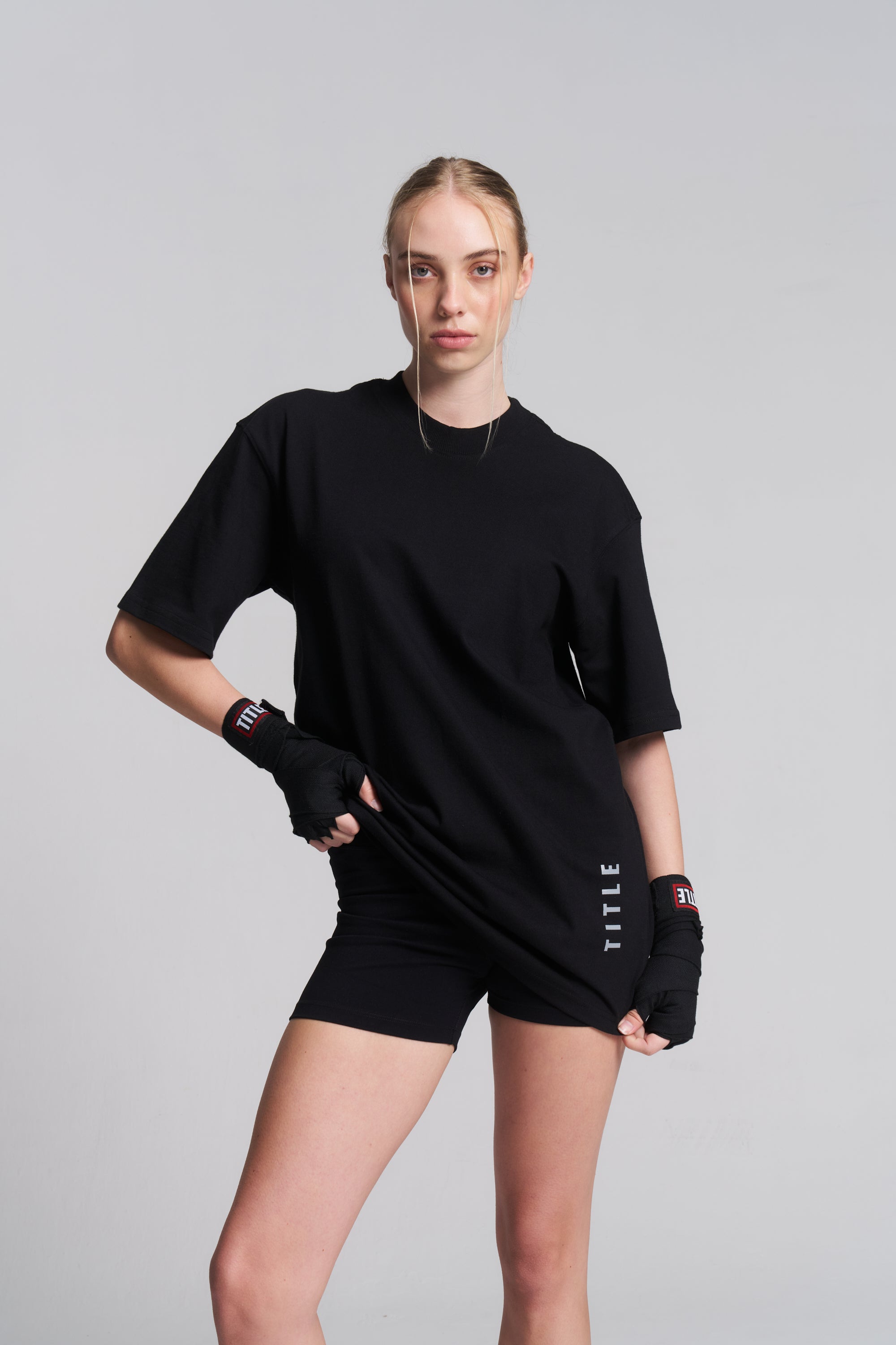 A woman modeling a black oversized short-sleeve T-shirt and matching black athletic shorts against a light gray studio background. She is wearing black fingerless gloves, and the shirt has white vertical text near the hem that reads 'TITLE'.