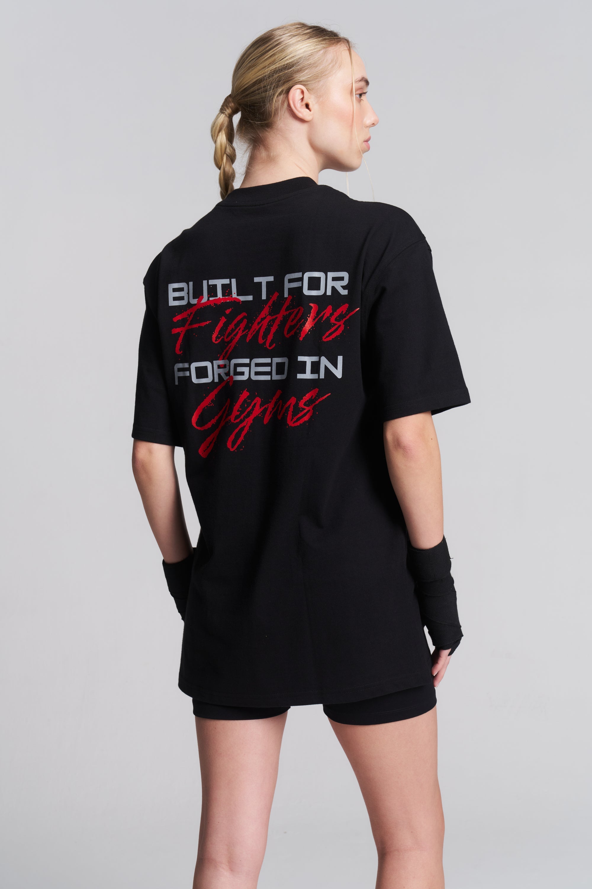 A woman shown from the back wearing a black oversized T-shirt with a large graphic text print on the back reading 'BUILT FOR Fighters FORGED IN Gyms' in white and red, paired with black shorts and wrist wraps, against a light gray studio background.