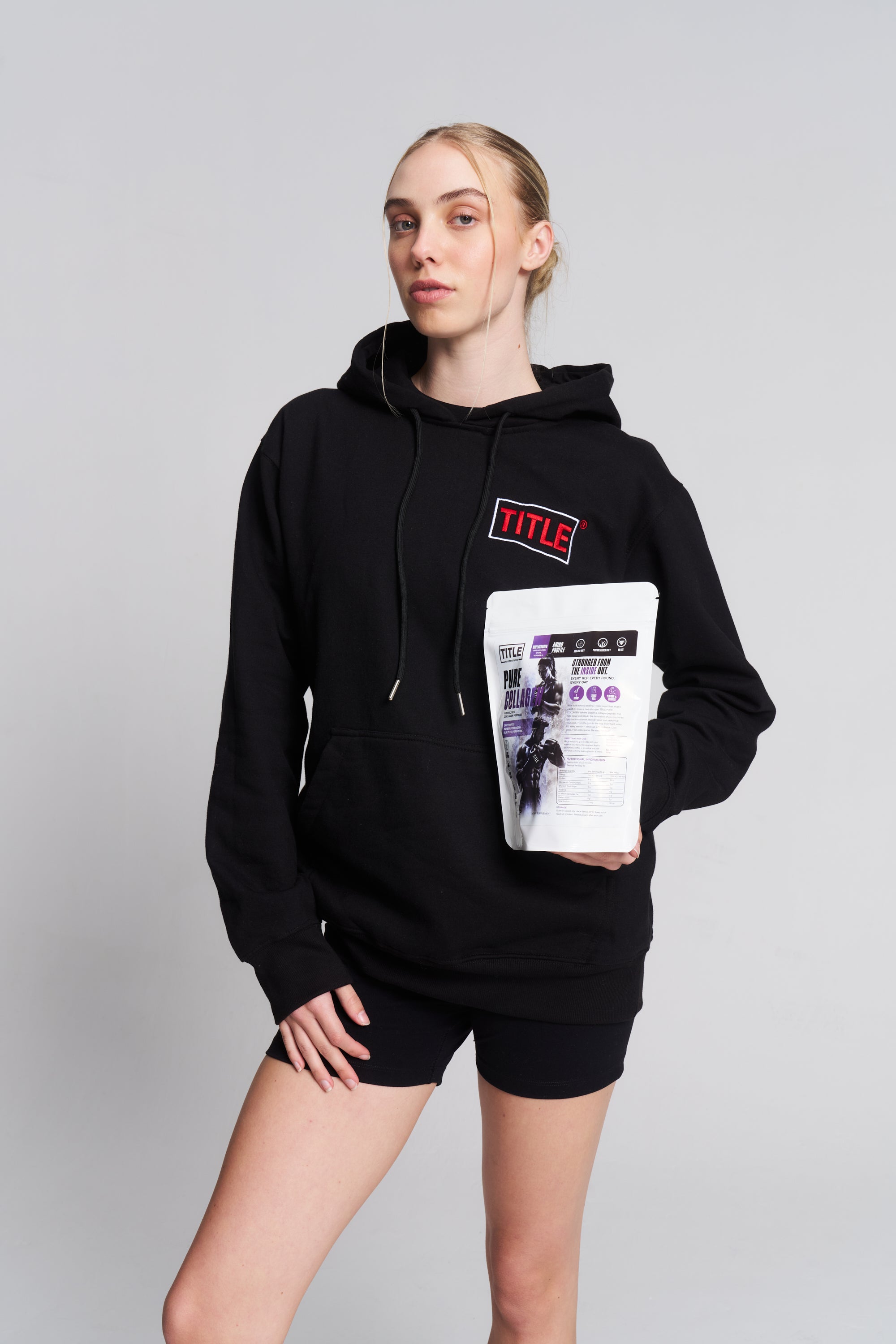 A woman wearing a black TITLE Boxing South Africa hoodie holding a bag of Pure Collagen hydrolysed bovine peptides.