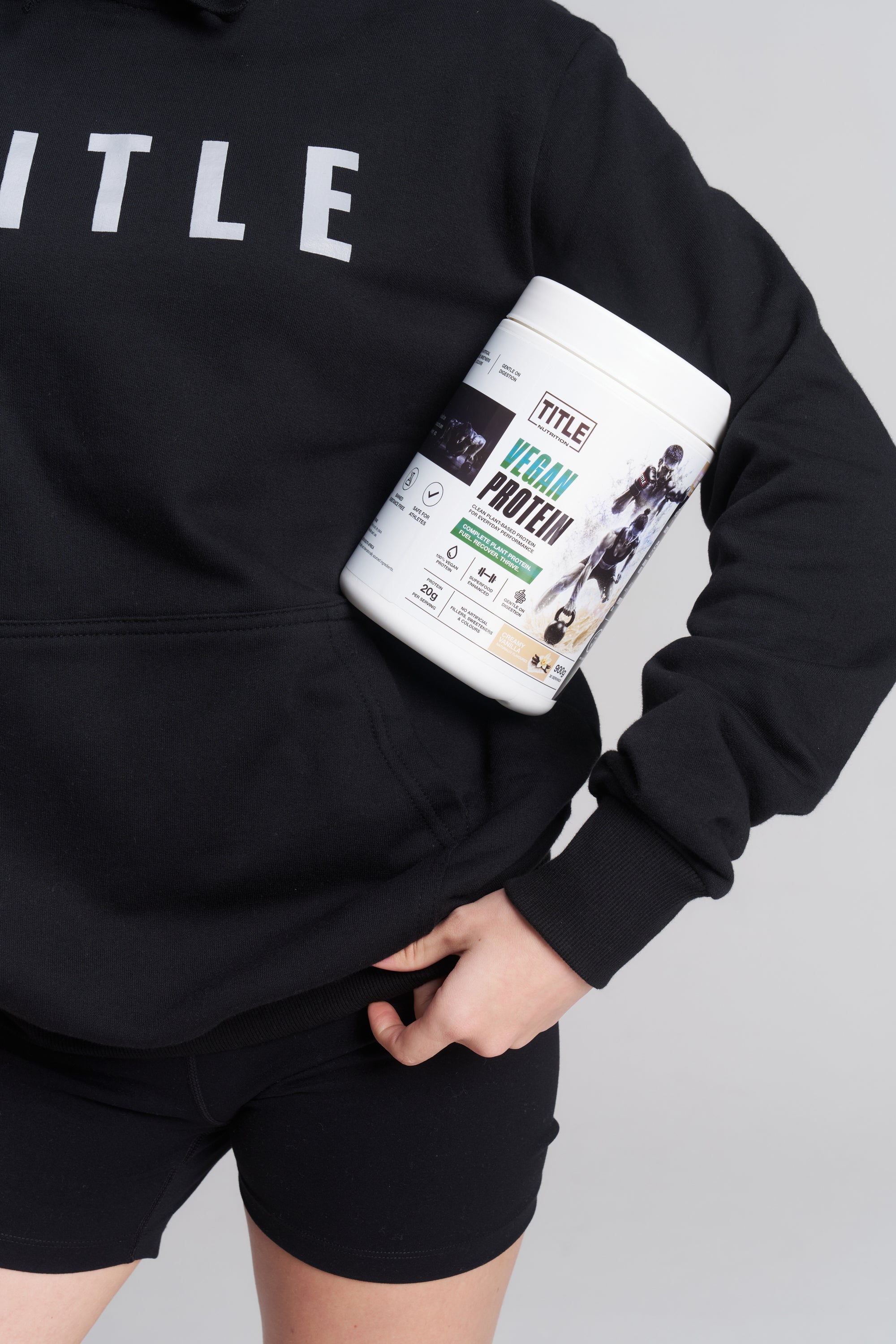 A person wearing a black TITLE Boxing South Africa hoodie holding a 900g tub of TITLE Vegan Protein powder.