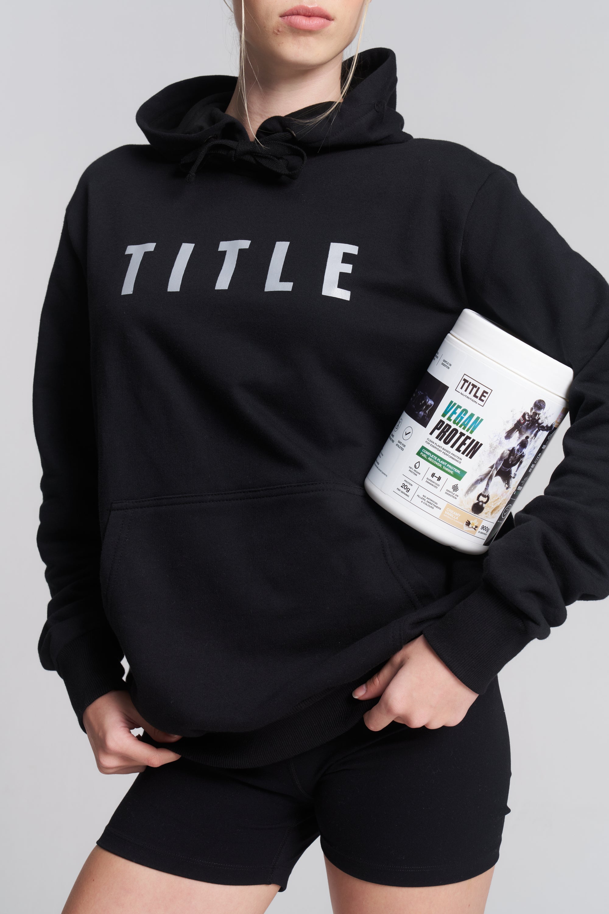 A person wearing a black TITLE Boxing South Africa hoodie holding a 900g tub of TITLE Vegan Protein powder.