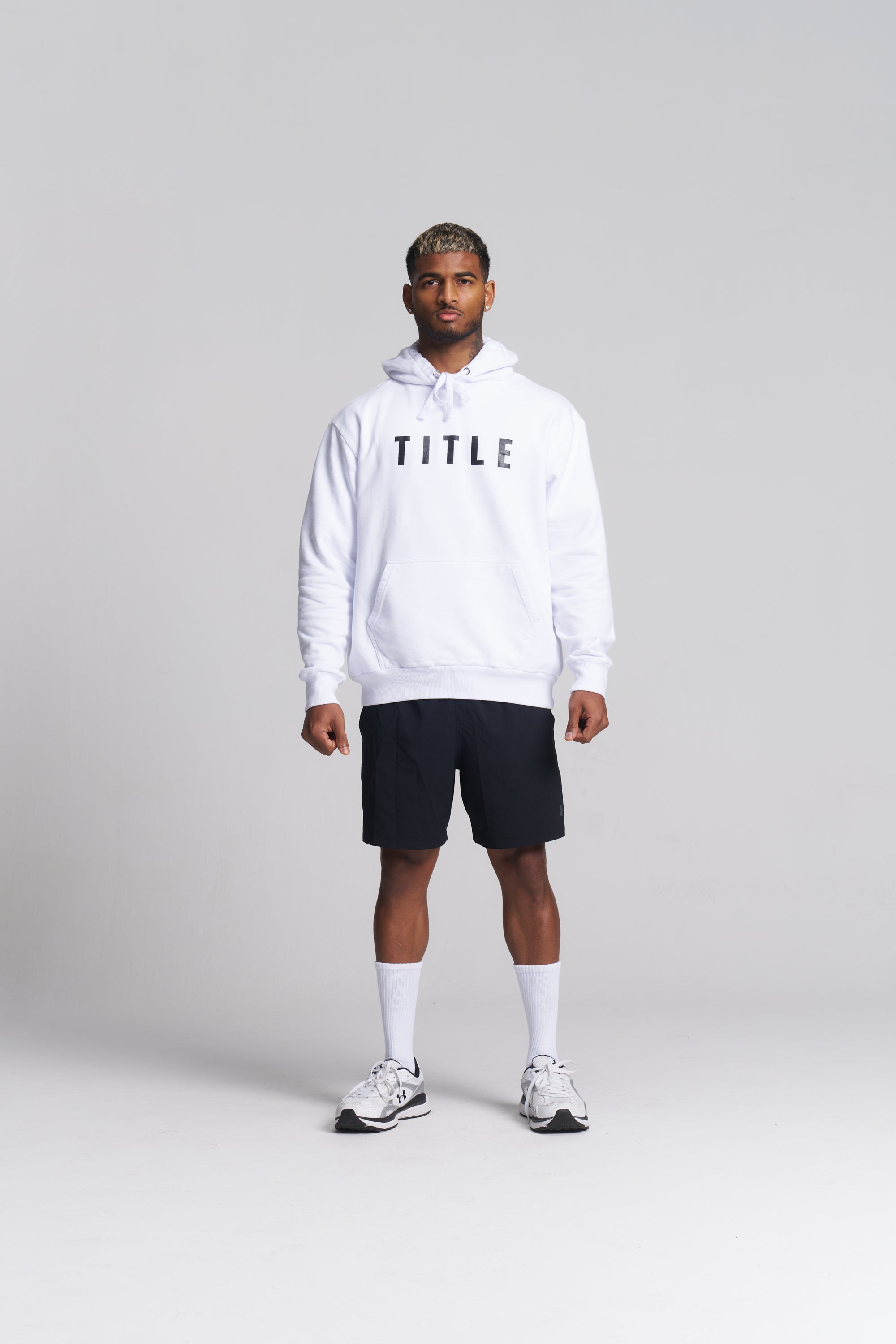 A man modeling a white pullover hoodie with 'TITLE' printed in black across the chest, paired with black athletic shorts, white socks, and white sneakers, against a light gray studio background.