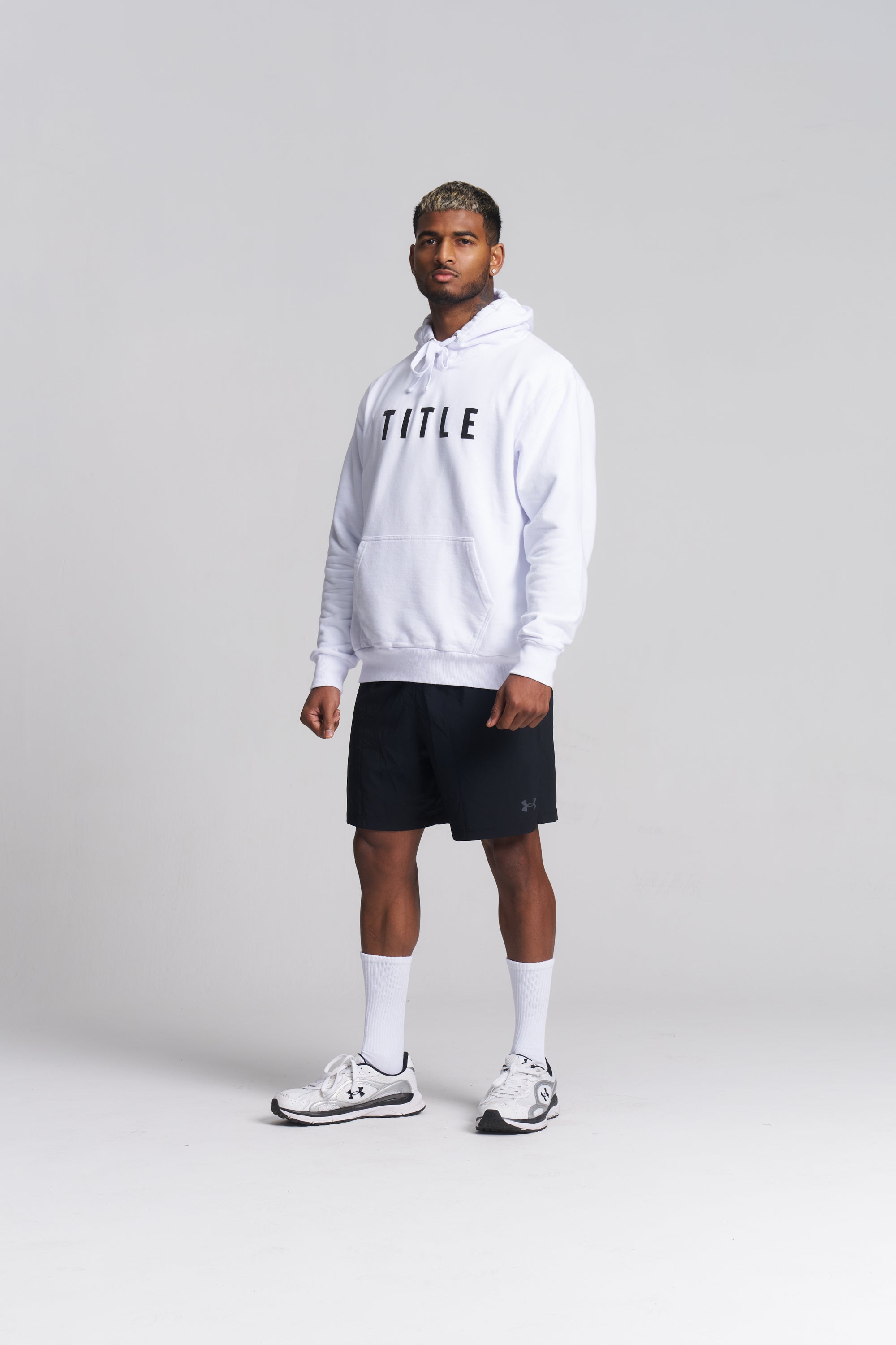 A male model stands in a studio wearing a white pullover hoodie with the word 'TITLE' printed in black across the chest, black athletic shorts, white socks, and white sneakers.