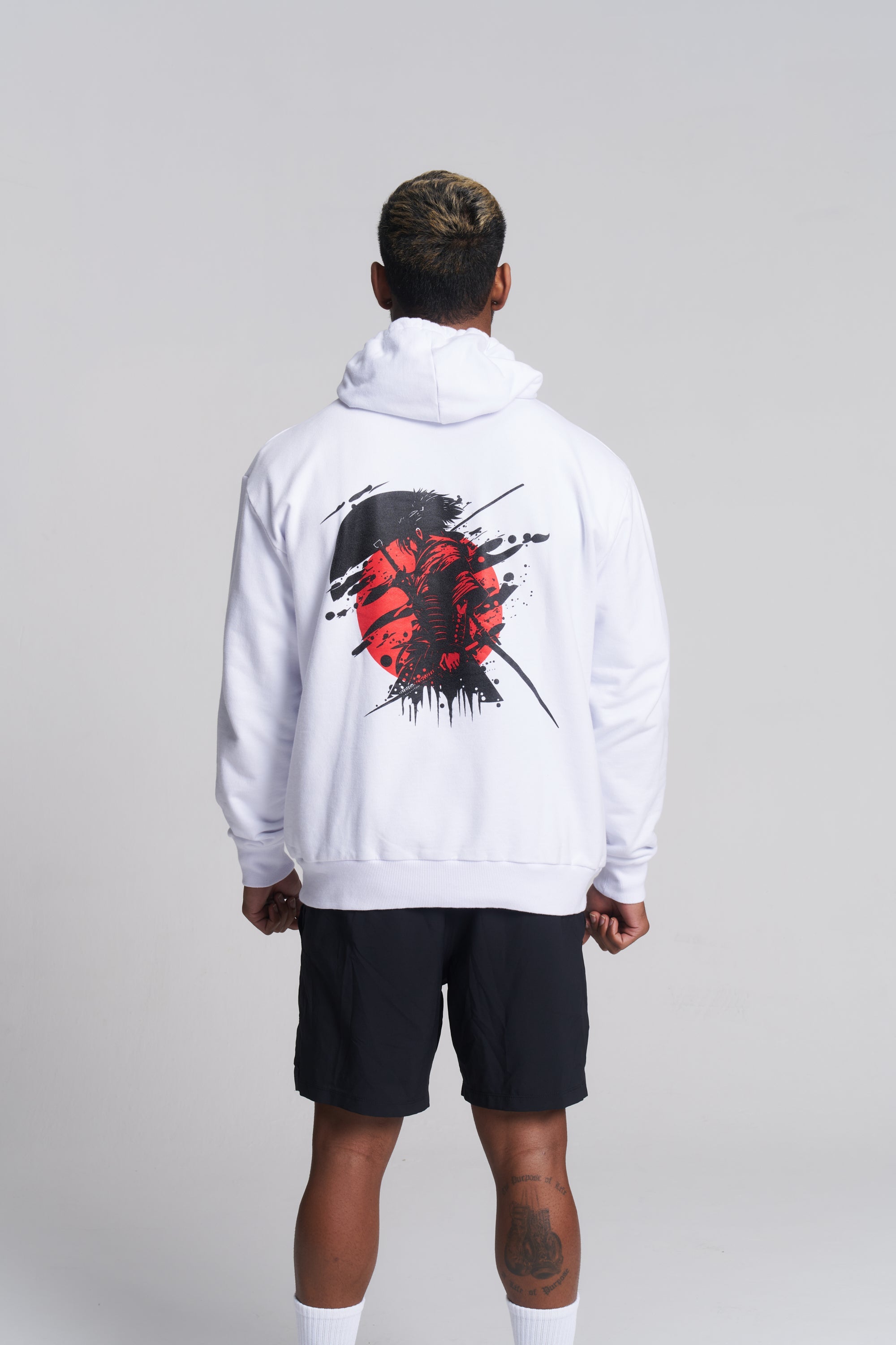 A man viewed from behind wearing a white pullover hoodie with a large red and black graphic print on the back, paired with black shorts and white socks, against a light gray studio background.