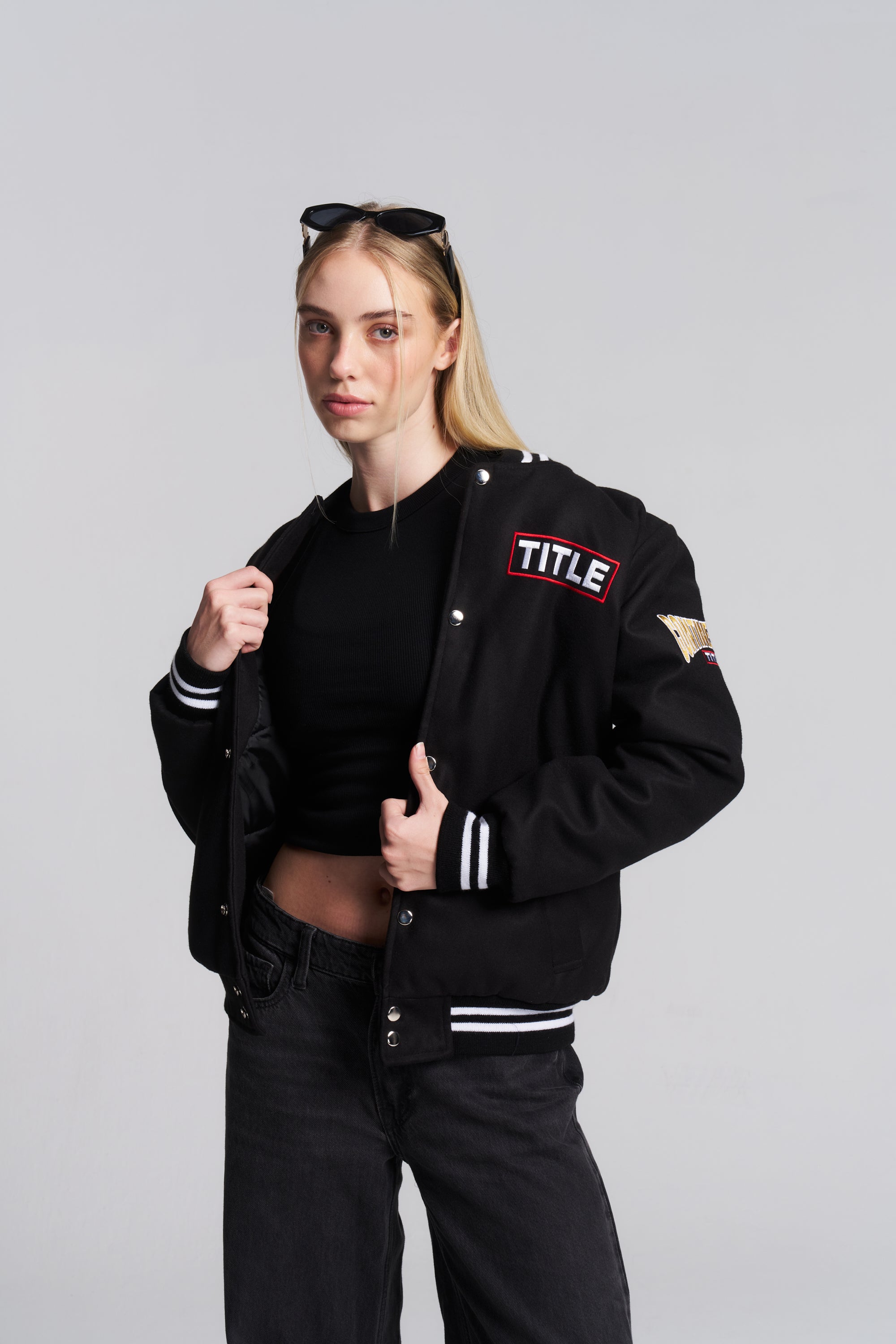 A blonde woman modeling a black varsity-style bomber jacket with white striped ribbed cuffs and hem, featuring a red and white 'TITLE' patch on the left chest, a gold patch on the right sleeve, and sunglasses on her head.