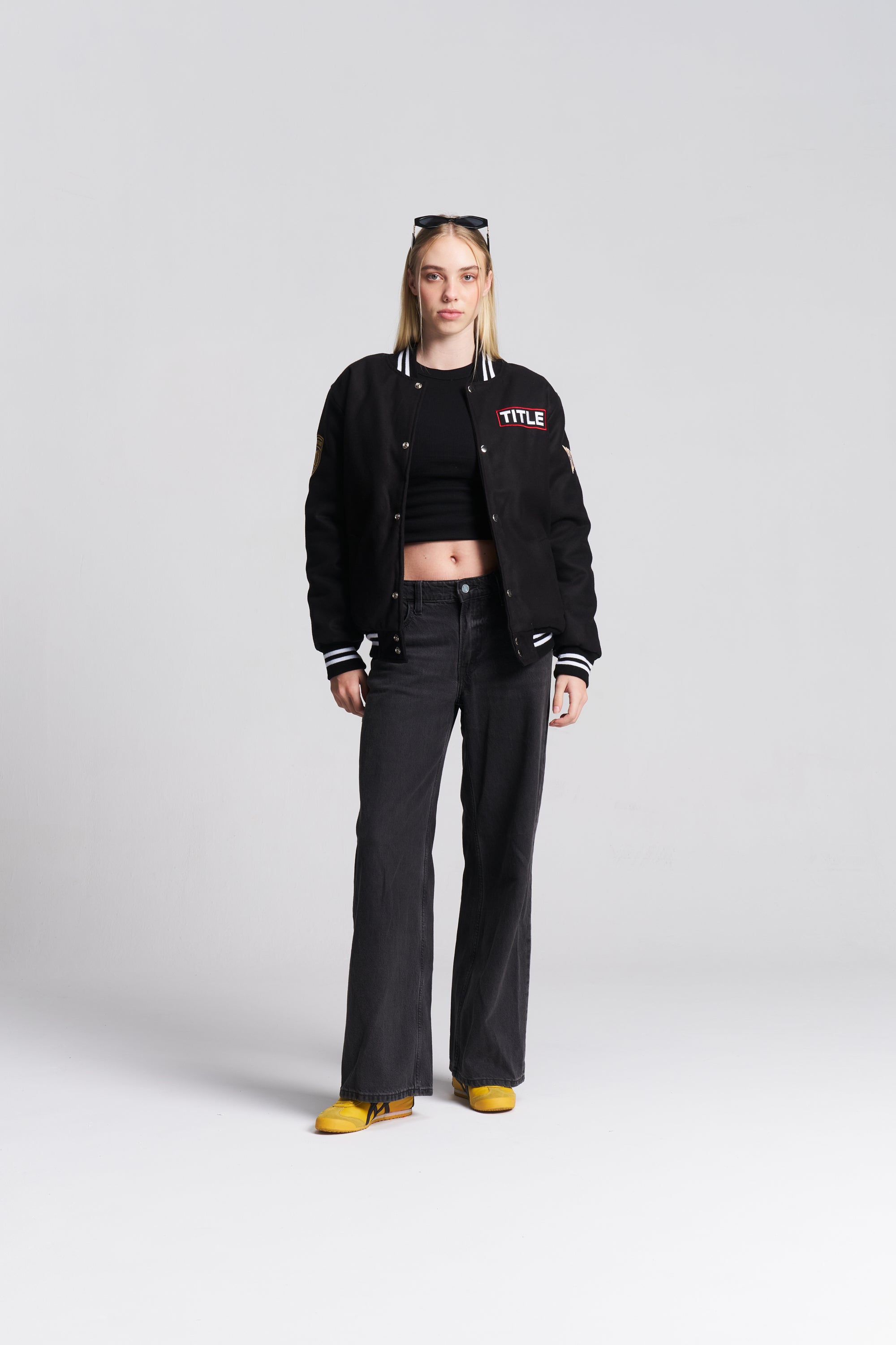 A model stands against a light gray studio background wearing a black varsity-style jacket with white and red lettering on the chest, black pants, and yellow shoes. The jacket has white striped ribbed trim and a white embroidered 'TITLE' logo on the left chest.