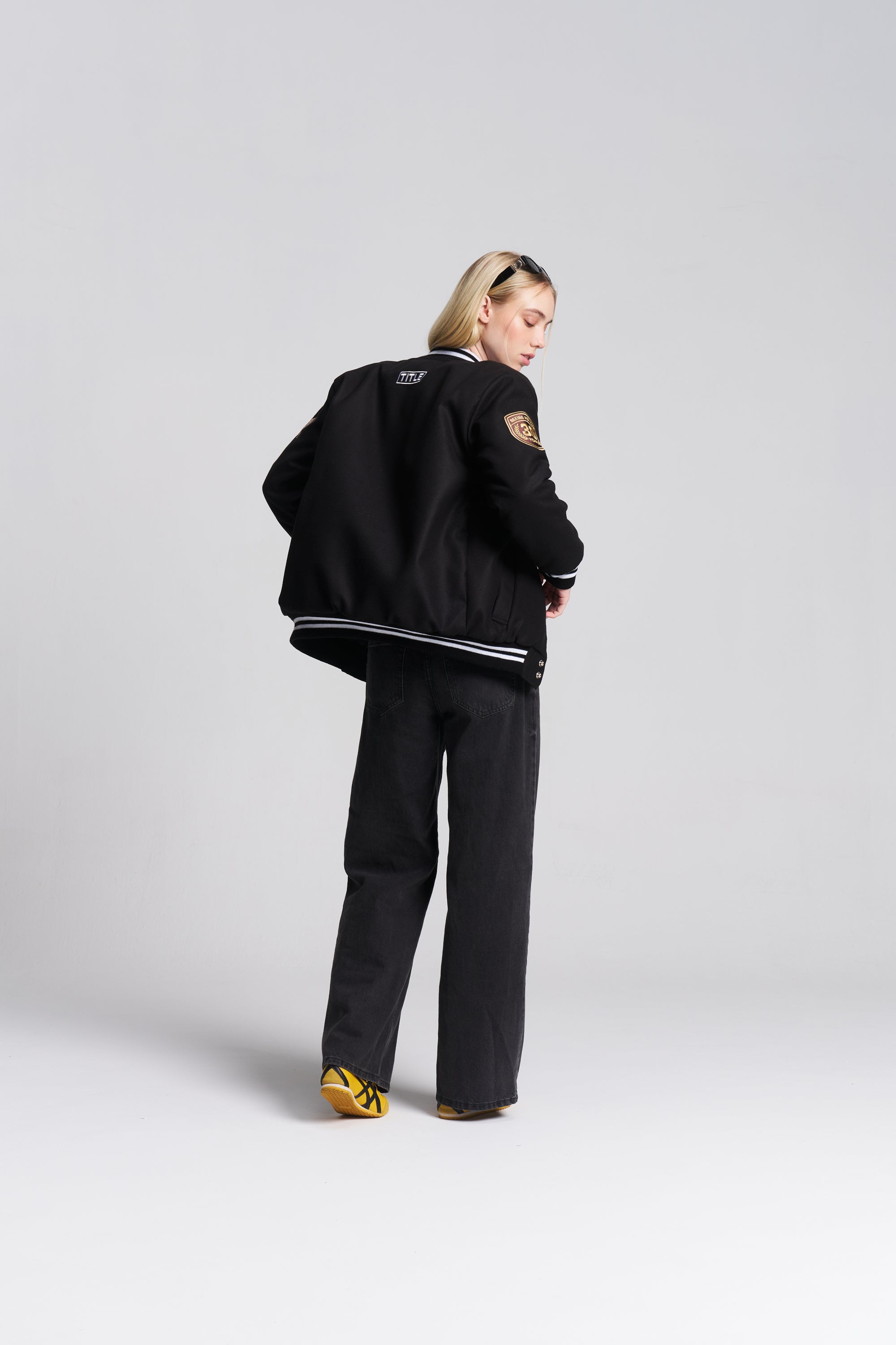 A model is shown from the back wearing a black varsity-style jacket with white stripes on the collar and cuffs, black wide-leg trousers, and yellow sneakers.