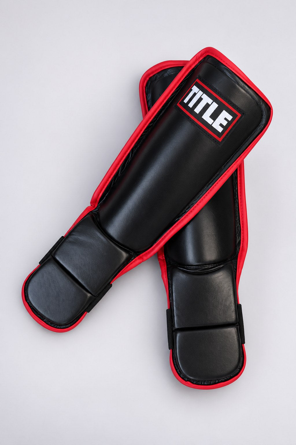 Pair of black leather training shin guards by TITLE laid crossed, showing the smooth shell and split instep pad