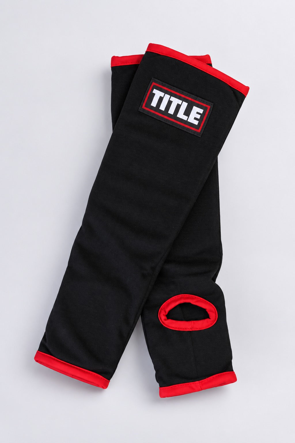 Pair of black slip-on MMA shin guards by TITLE laid crossed, showing red binding and the heel opening