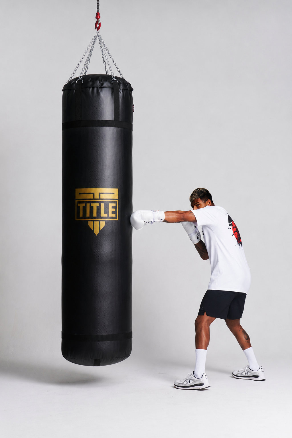 A man in a white t-shirt, black shorts, and white boxing gloves punches a black heavy punching bag hanging from chains. The bag features a gold and black TITLE logo.